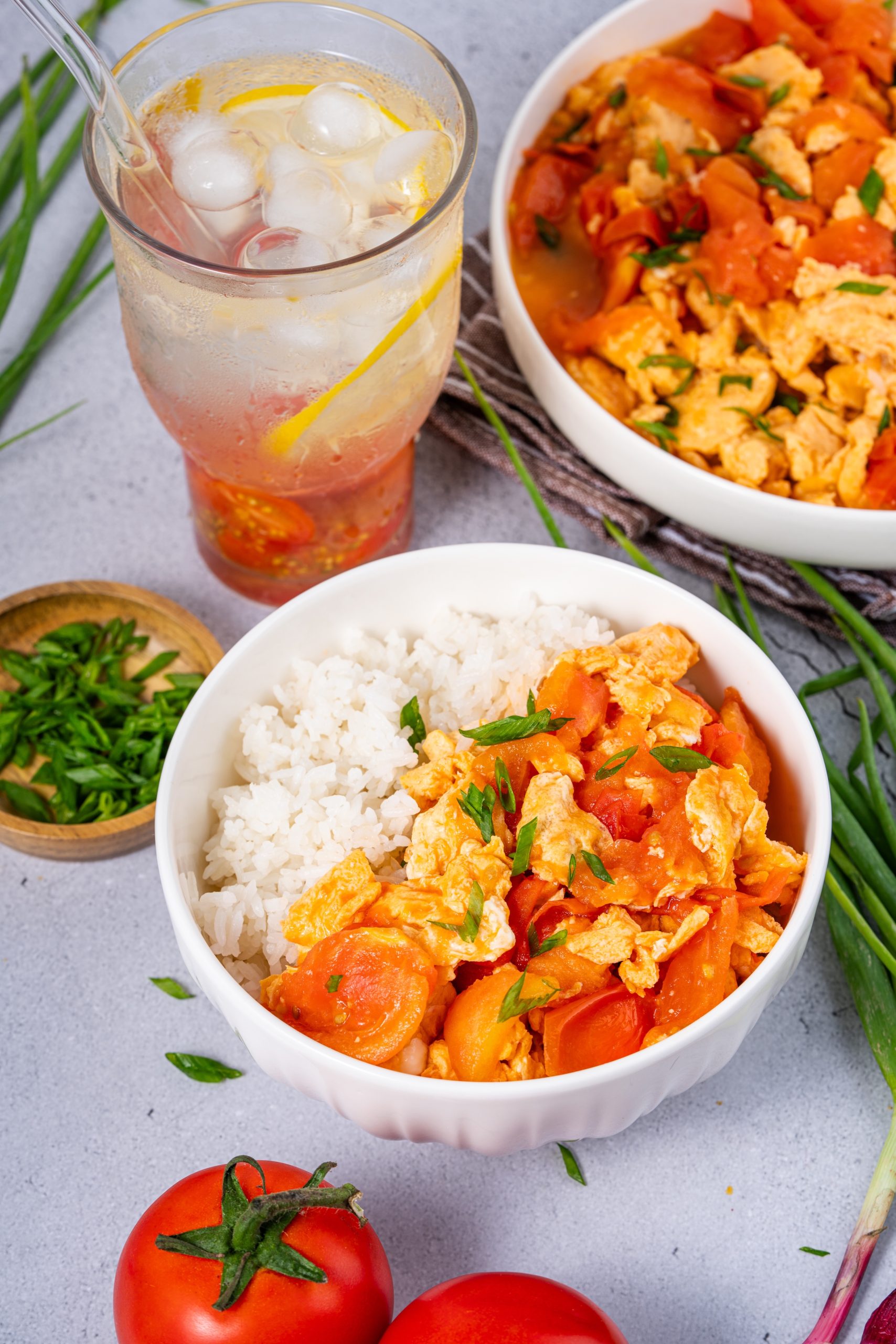 Bowl of rice topped with Stir Fried Tomato and Egg Recipe, styled with tomatoes, scallions, and a refreshing lemon and tomato iced drink.