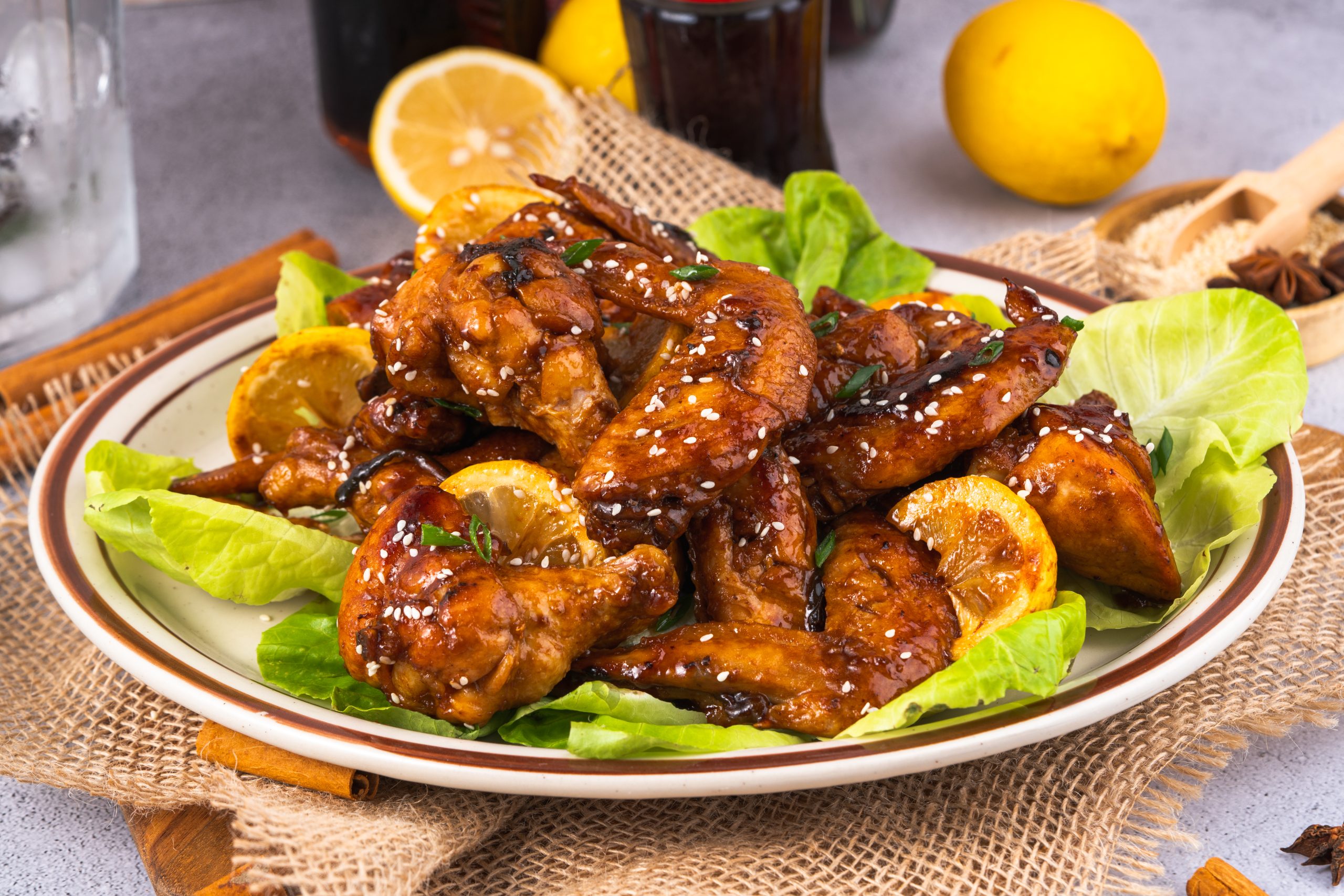 Wide plate of Coca Cola Chicken Wings with glazed lemon slices and fresh lettuce, styled with lemons, Coke, and spices.