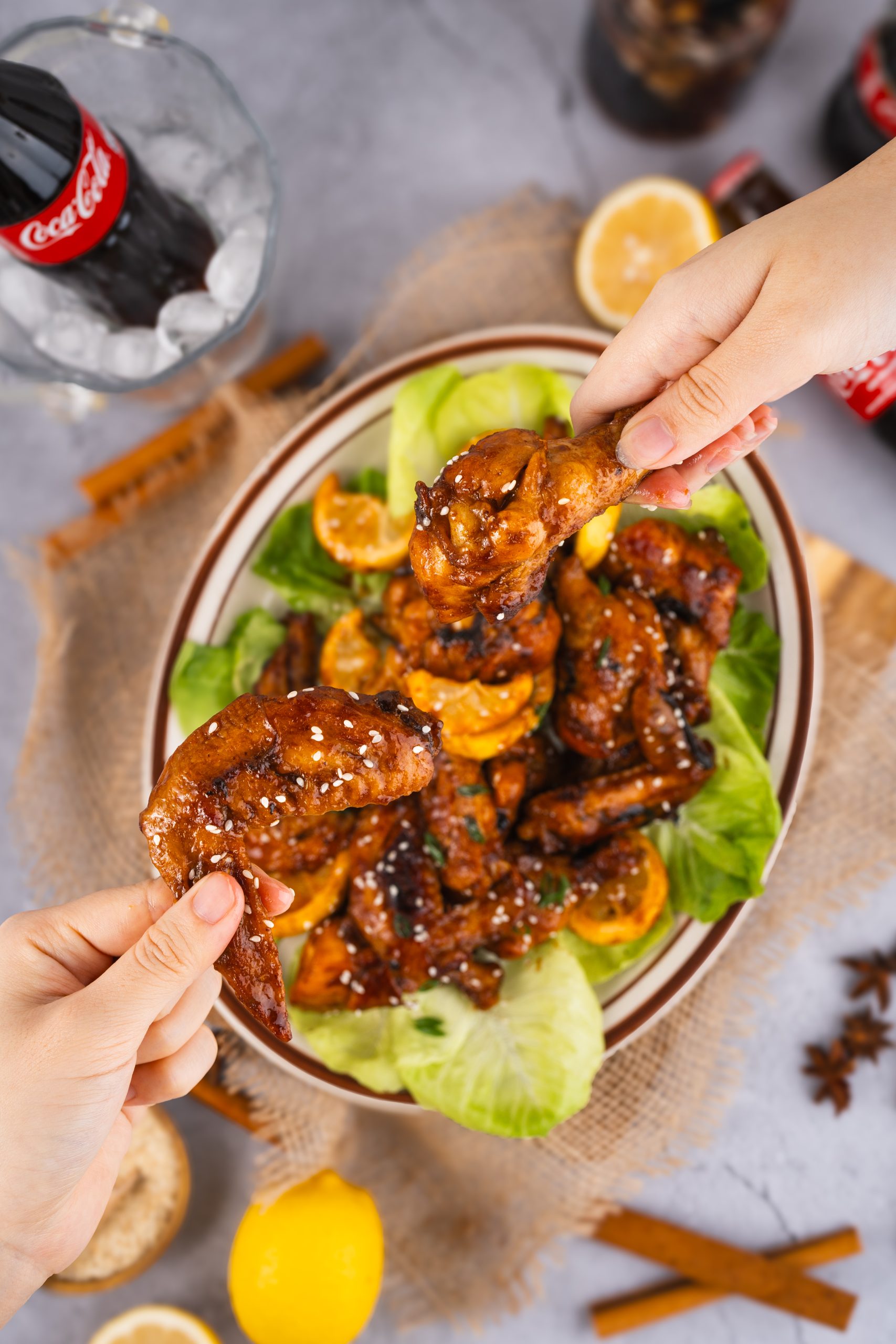 Two hands holding Coca Cola Chicken Wings over a plate, ready to eat, with a cold Coke and citrus garnish in background.