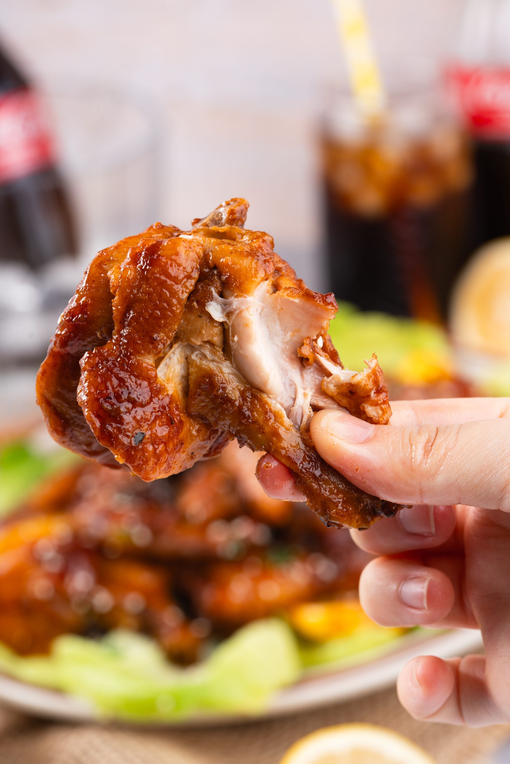 Close-up of a hand holding a juicy Coca Cola Chicken Wing with a bite taken out, showing the tender interior and glossy glaze.