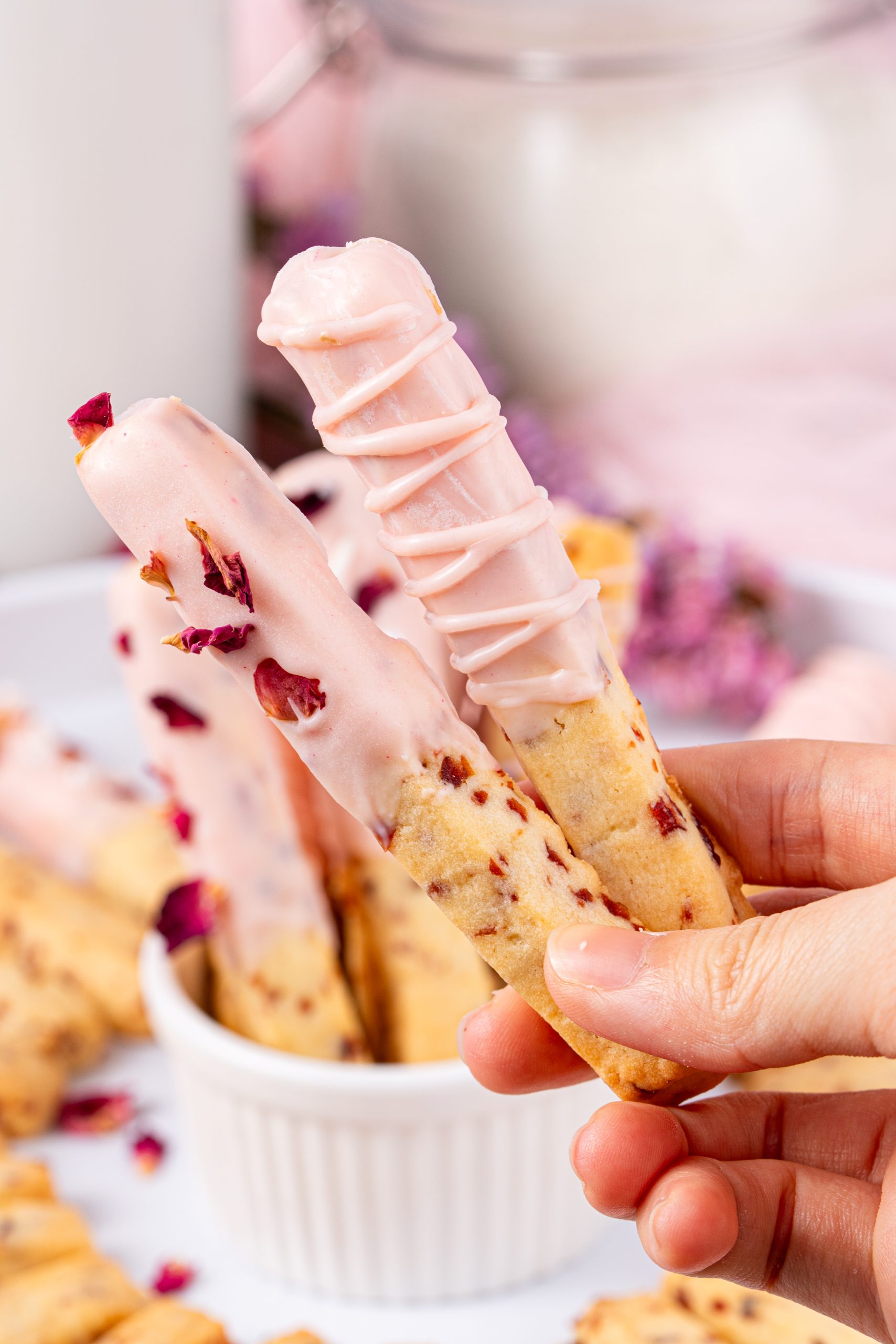 Hand holding two Cranberry Cookies dipped in pink-tinted white chocolate, decorated with rose petals and drizzle.
