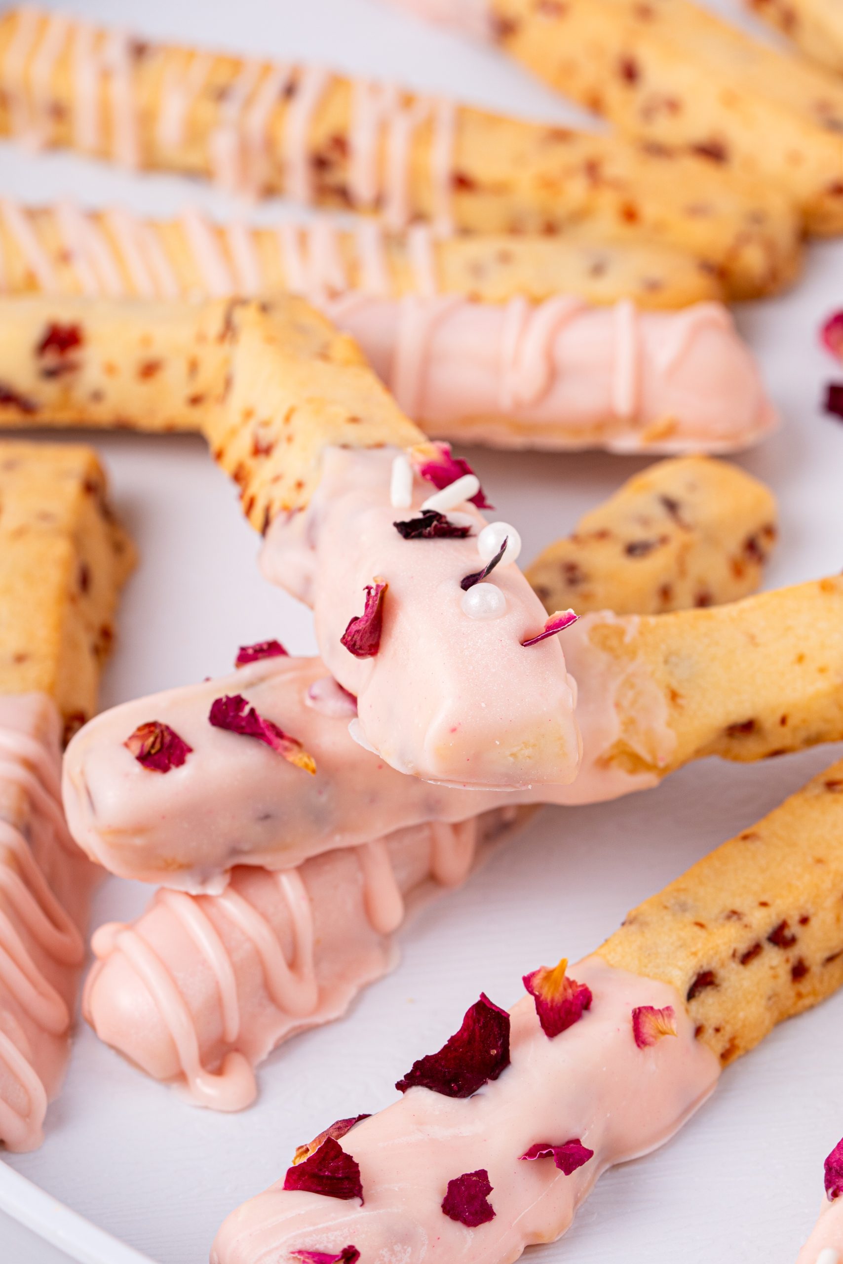Cranberry cookie sticks dipped in white chocolate and decorated with rose petals and sprinkles on a white plate.