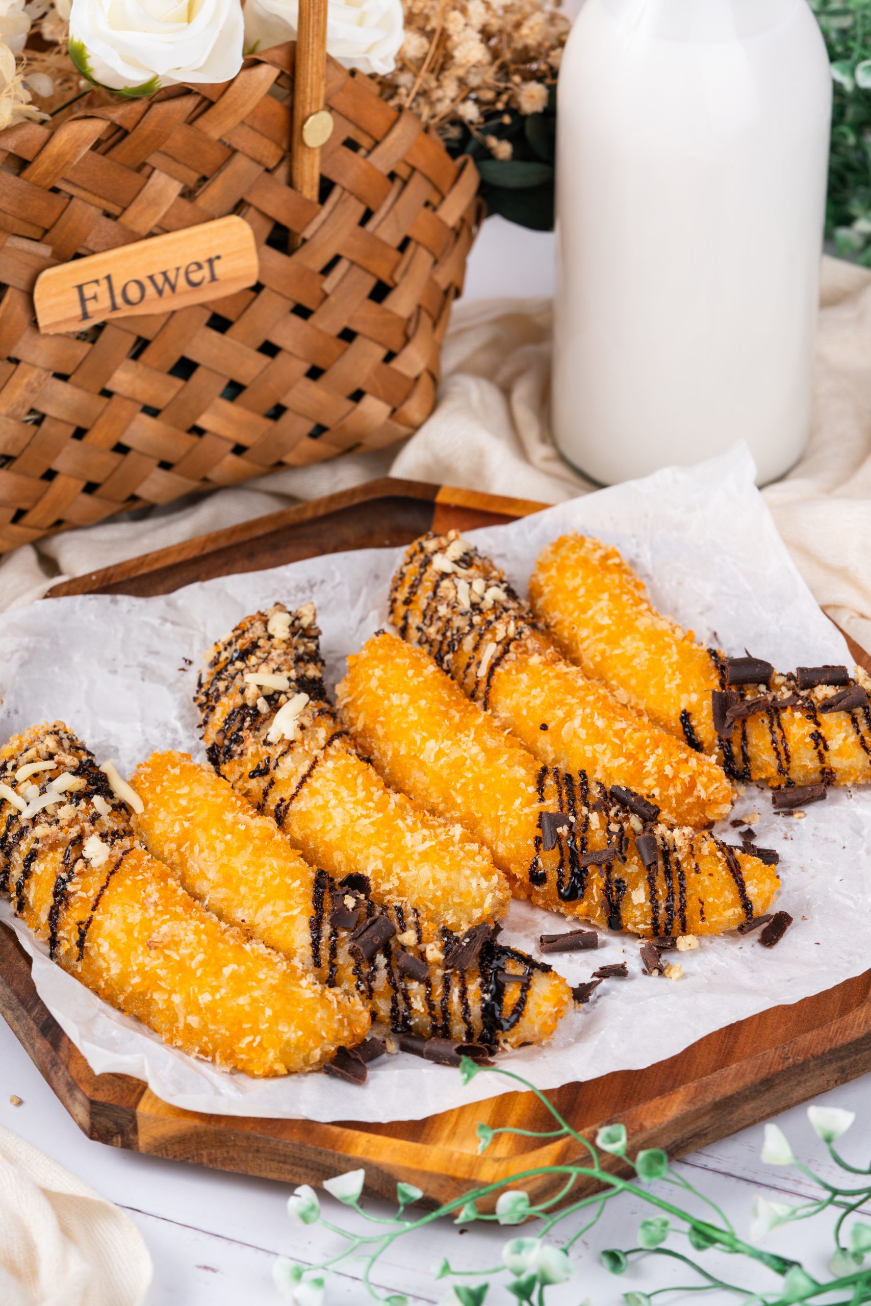 Crispy banana fritters recipe topped with chocolate drizzle and shredded cheese, served on parchment paper over a wooden tray beside a basket of flowers and a bottle of milk.