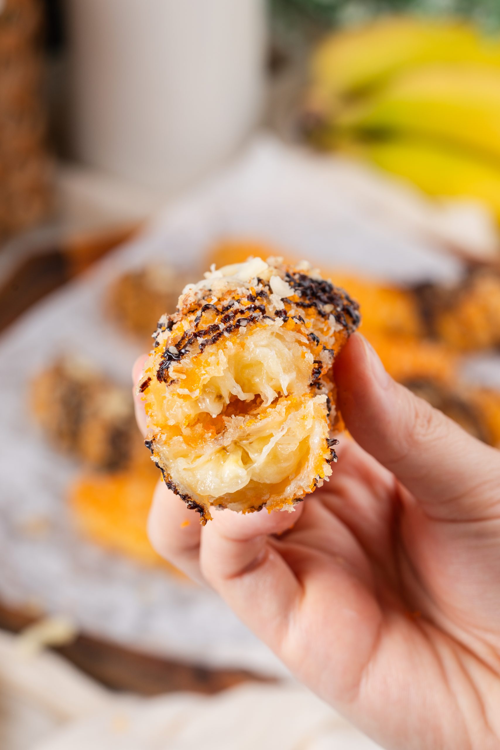 Close-up of a bitten banana fritter recipe showing its soft banana interior and crispy coating, topped with chocolate and nuts.