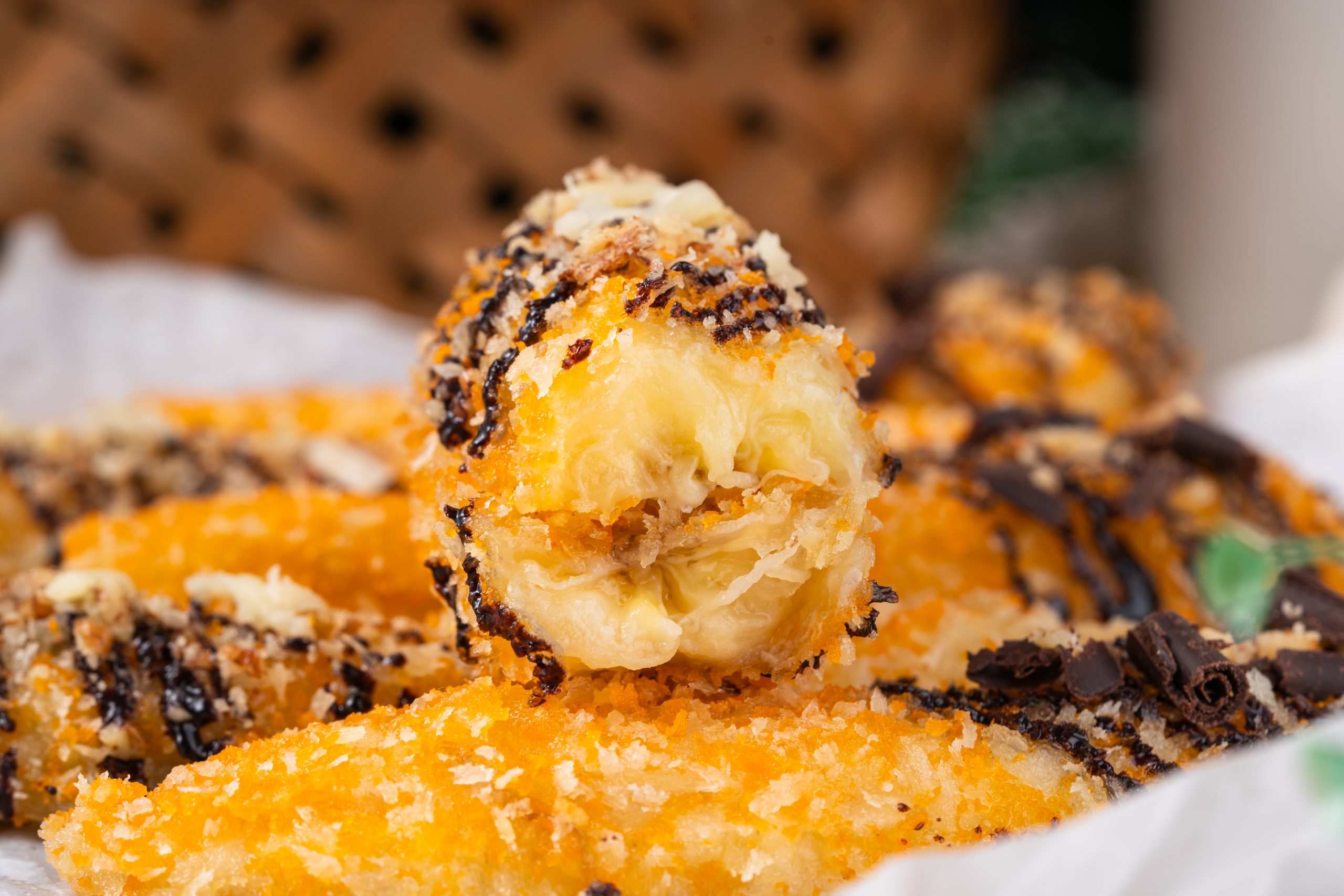Macro view of a banana fritter recipe showing its golden, crunchy coating and creamy banana filling, drizzled with chocolate and surrounded by more fritters on parchment paper.