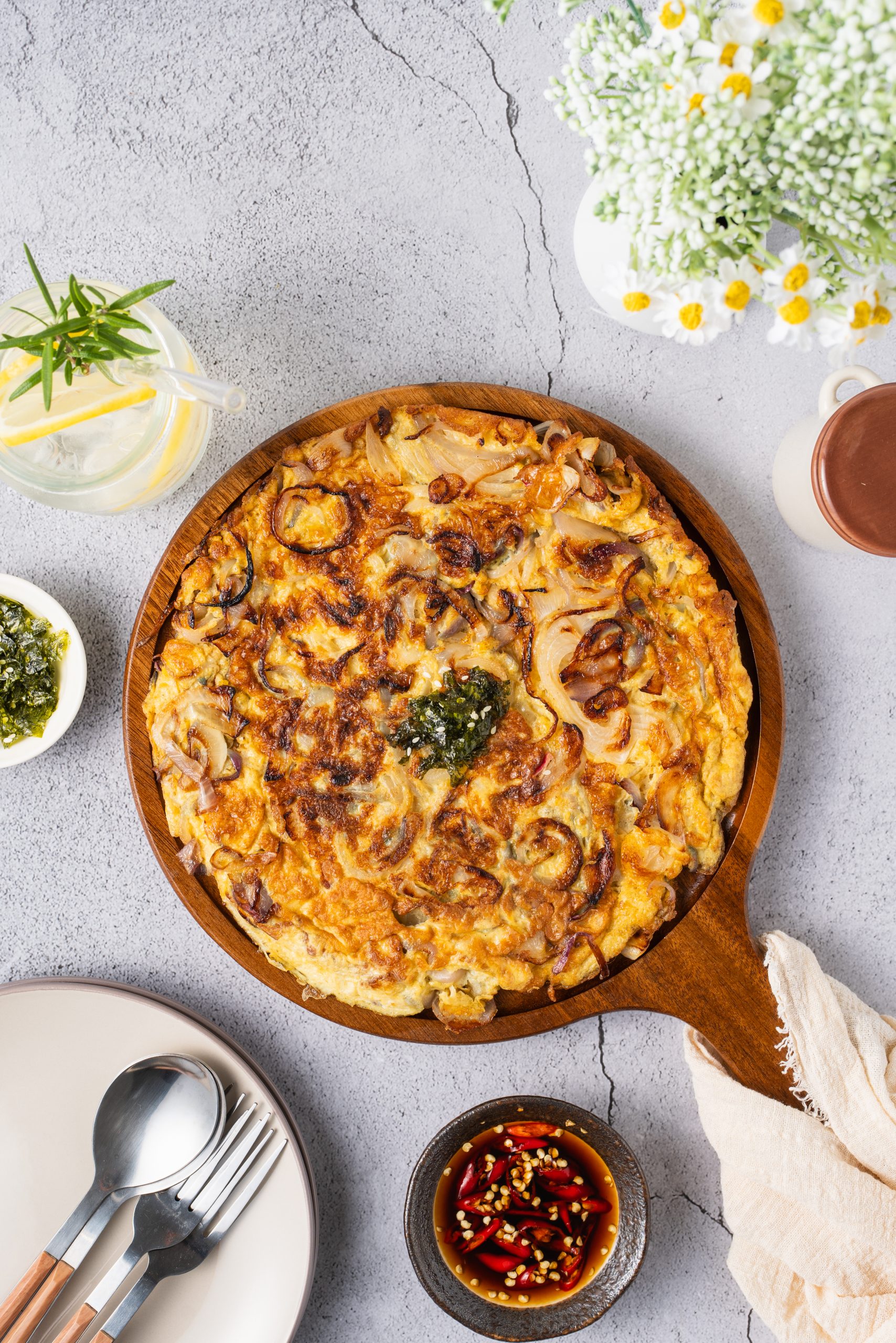 A delicious Chinese onion omelette recipe served on a wooden plate with dipping sauce, lemon water, and flowers on a gray tabletop.