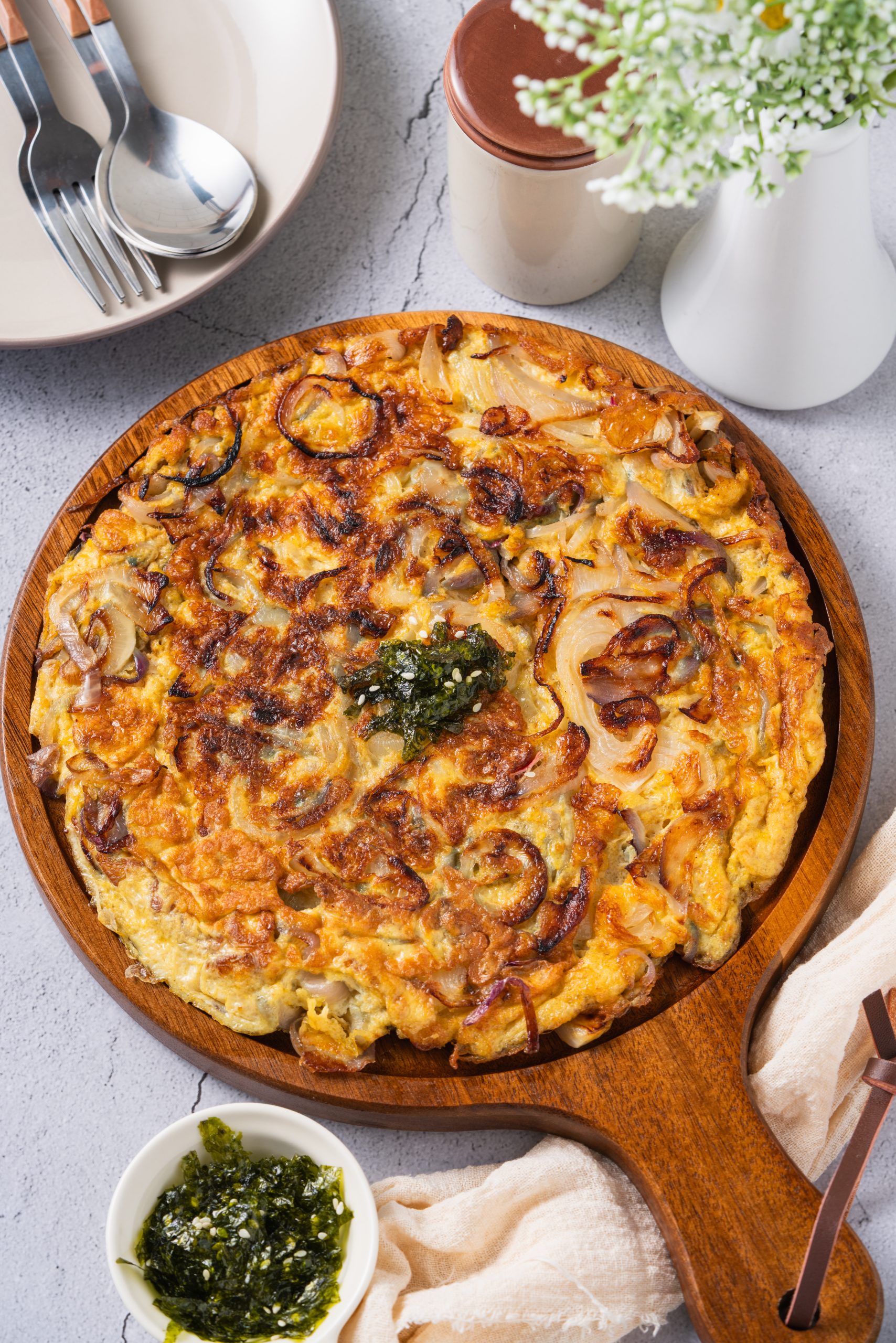 Overhead view of a golden-brown Onion omelette served on a wooden board, surrounded by soy dipping sauce, fresh flowers, and a glass of lemon water on a textured grey table.