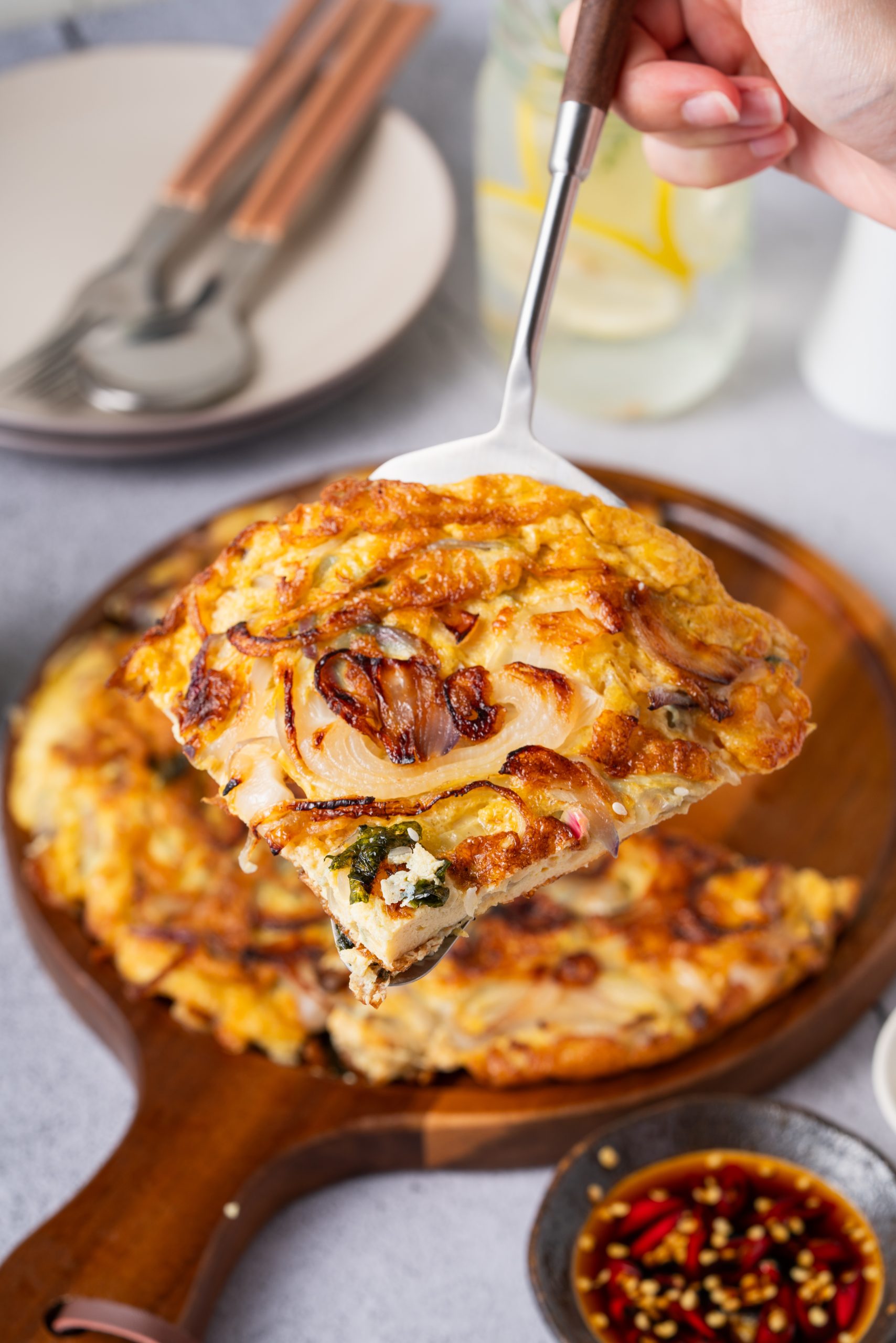A beautifully cooked Chinese onion omelette recipe served on a wooden board, garnished with seaweed and sesame seeds, placed beside a small bowl of sauce, tableware, and a white vase with flowers on a light gray surfa