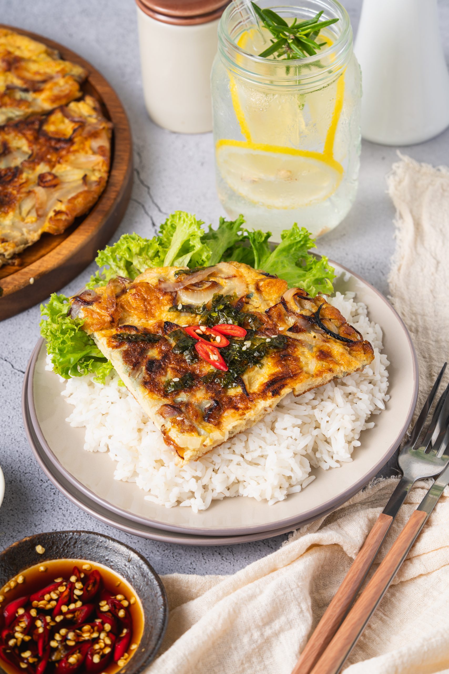A plate of Chinese onion omelette on rice with lettuce, a glass of lemon water, and sauce on the side; the omelette is garnished with seaweed and chili.