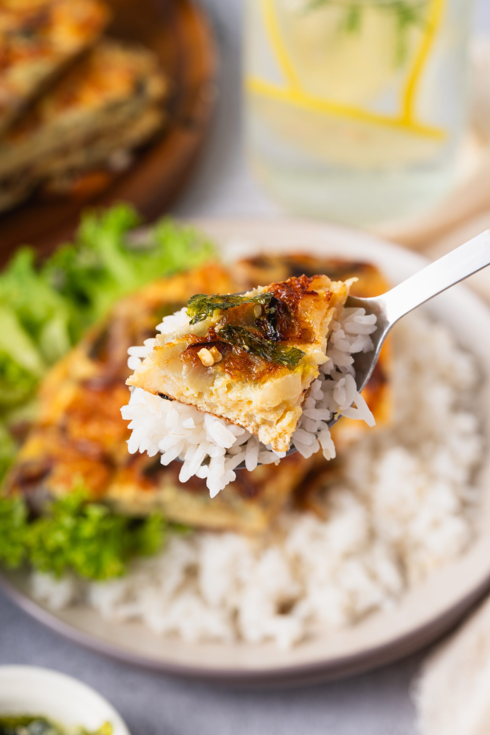A forkful of Chinese onion omelette recipe with rice, topped with caramelized onions and seaweed, held above a plate of rice and lettuce.