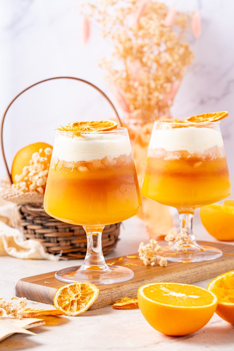 Two glasses of orange Earl Grey tea topped with thick cheese foam and garnished with dried orange slices, styled with fresh oranges and soft floral accents.