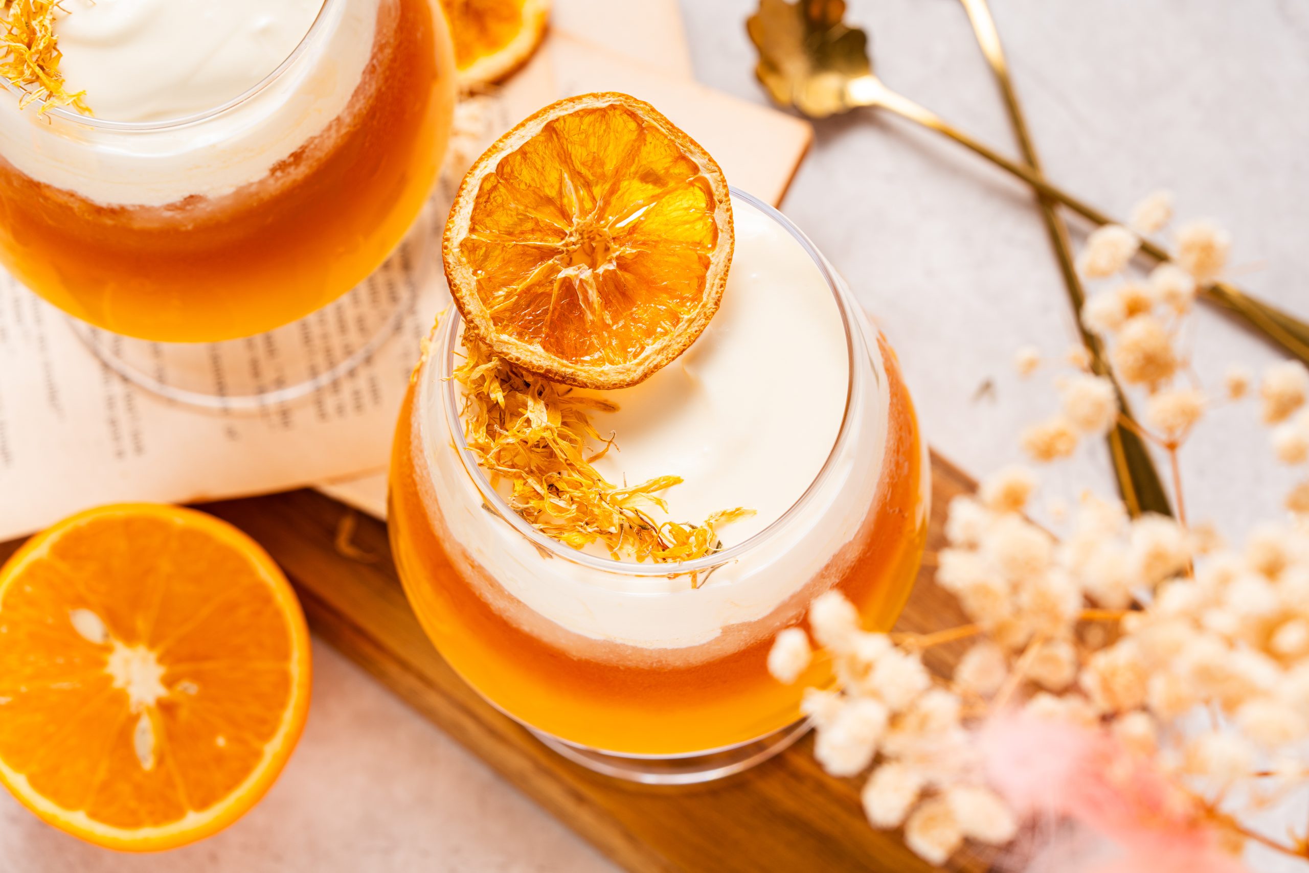 Top view of two glasses of orange Earl Grey tea with cheese foam, garnished with dried orange slices, styled alongside open book pages and scattered dried flowers.