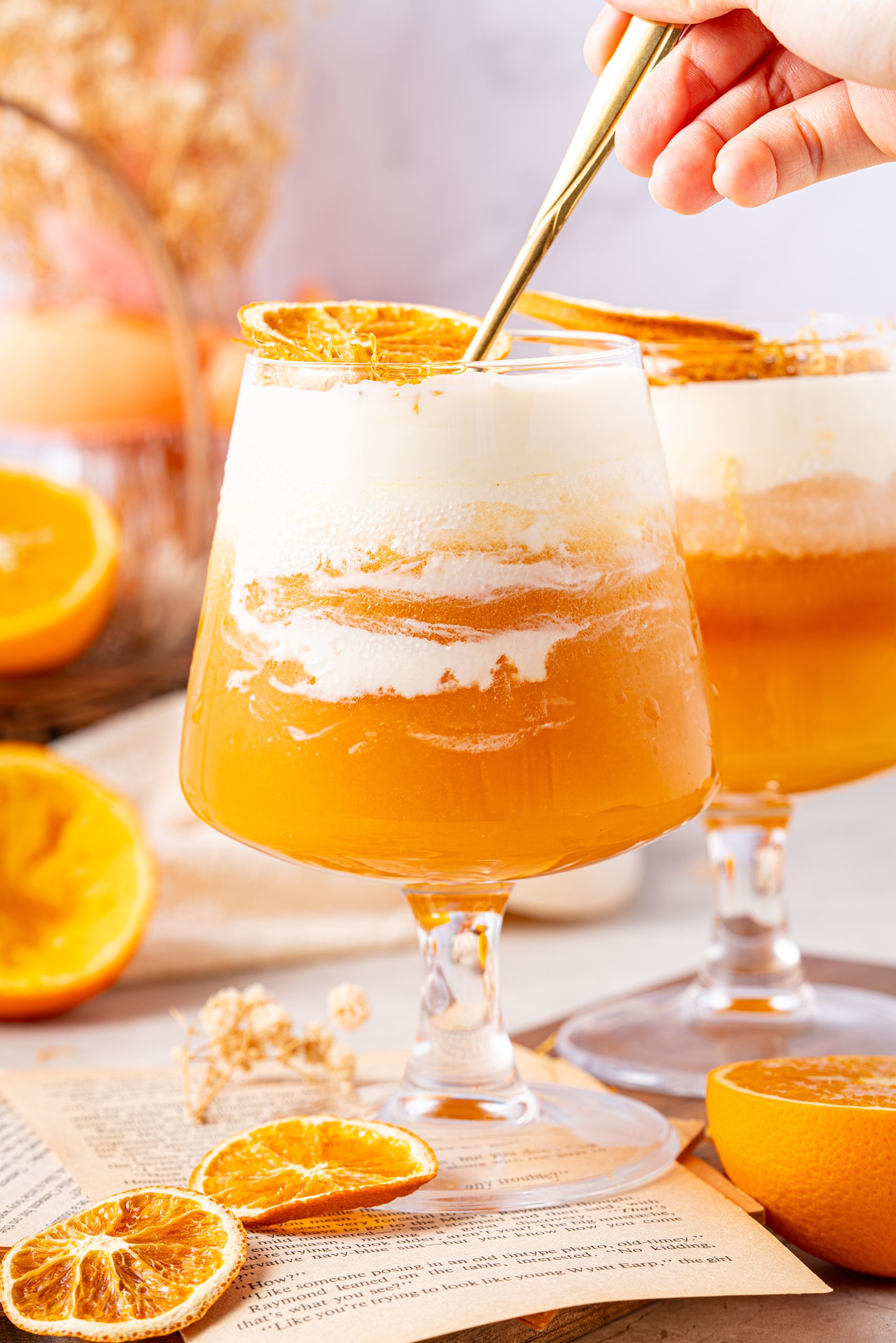 Hand stirring a glass of orange tea topped with thick cheese foam, garnished with a dried orange slice, with another layered tea drink and citrus decor in the background.