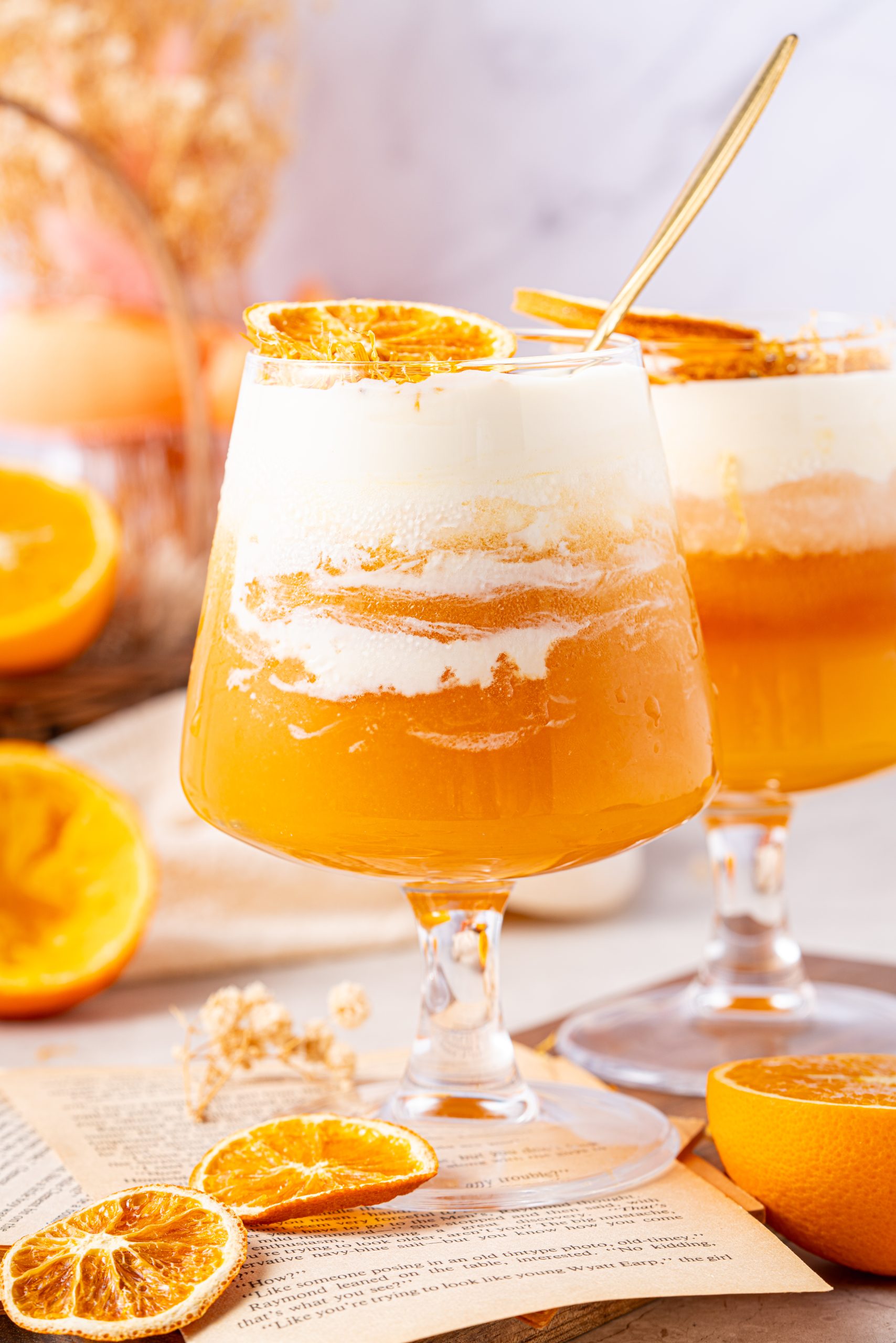 Two chilled glasses of orange tea topped with frothy cheese foam, one with a spoon inside, styled with halved oranges, dried citrus, and floral decor.