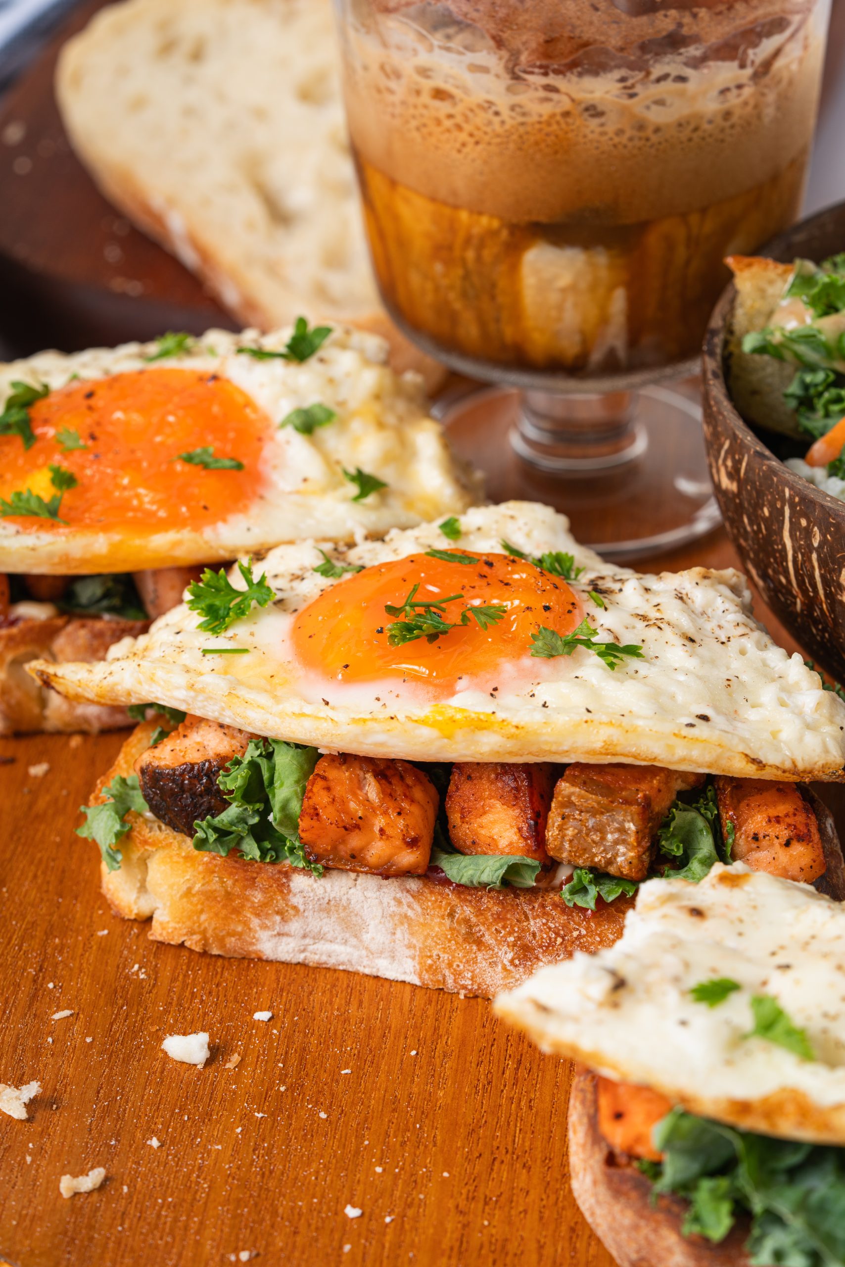Close-up of gourmet Salmon Sandwiches with crispy sourdough bread, golden fried eggs, fresh kale, and tender salmon cubes, served with iced coffee and salad.