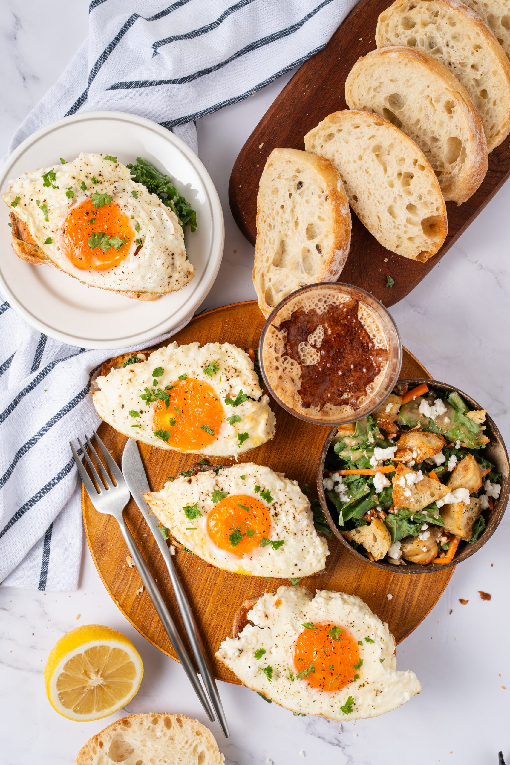 Overhead view of freshly made Salmon Sandwiches topped with sunny-side-up eggs, served with rustic bread slices, salad, and a frothy iced coffee.