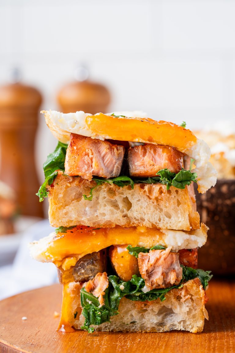 Stacked Salmon Sandwiches cut in half to show layers of juicy salmon, leafy greens, and fried eggs on fluffy sourdough bread.