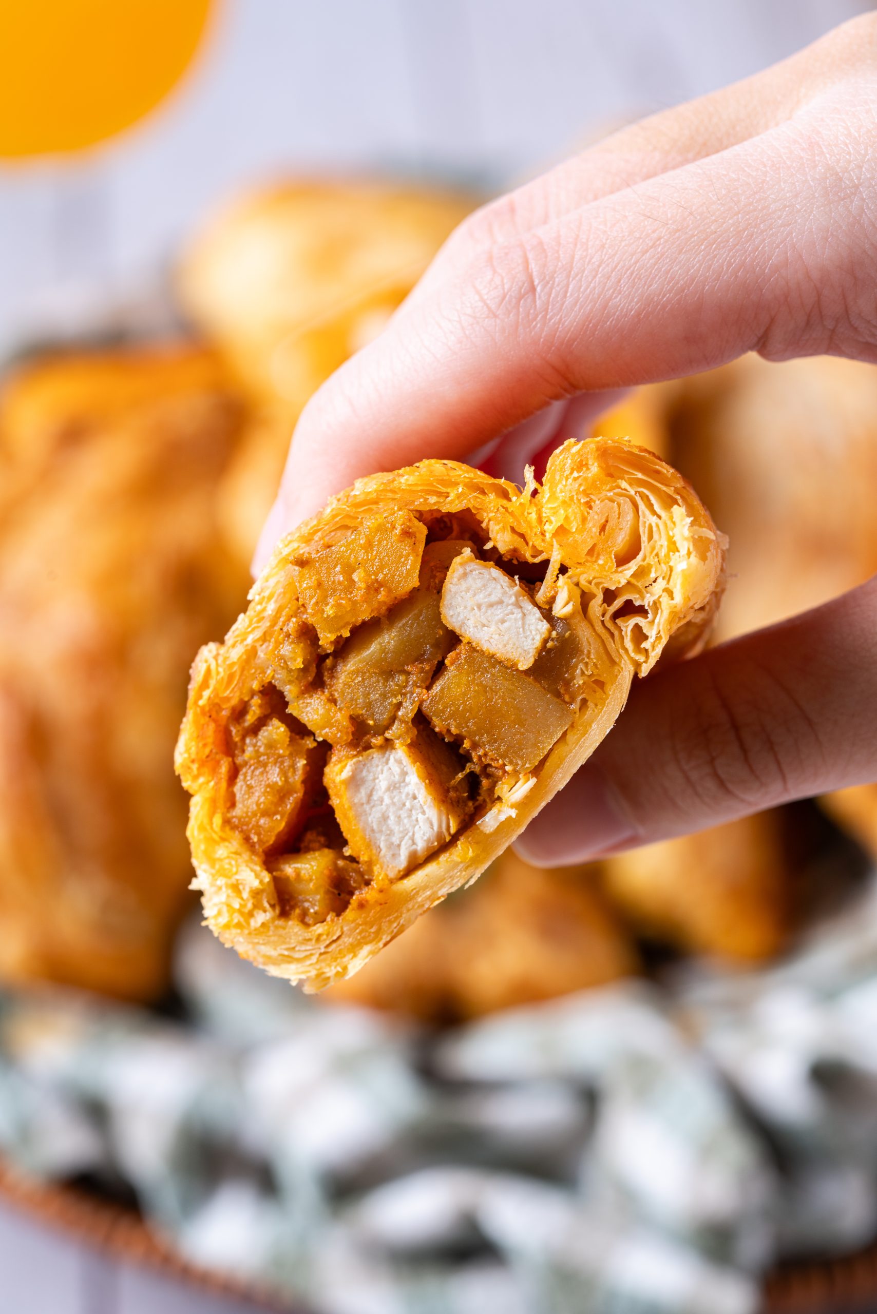 A hand holding a freshly baked Curry Puff, showing its golden flaky crust and savory filling of curried potatoes and chicken cubes.