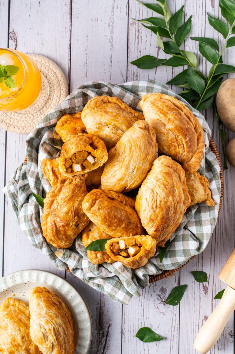 A basket of golden, flaky curry puffs filled with spiced potato and chicken, displayed on a checkered cloth. One puff is cut open to show the savory filling. The scene includes a glass of orange drink with ice, fresh curry leaves, and whole potatoes on a rustic wooden surface.
