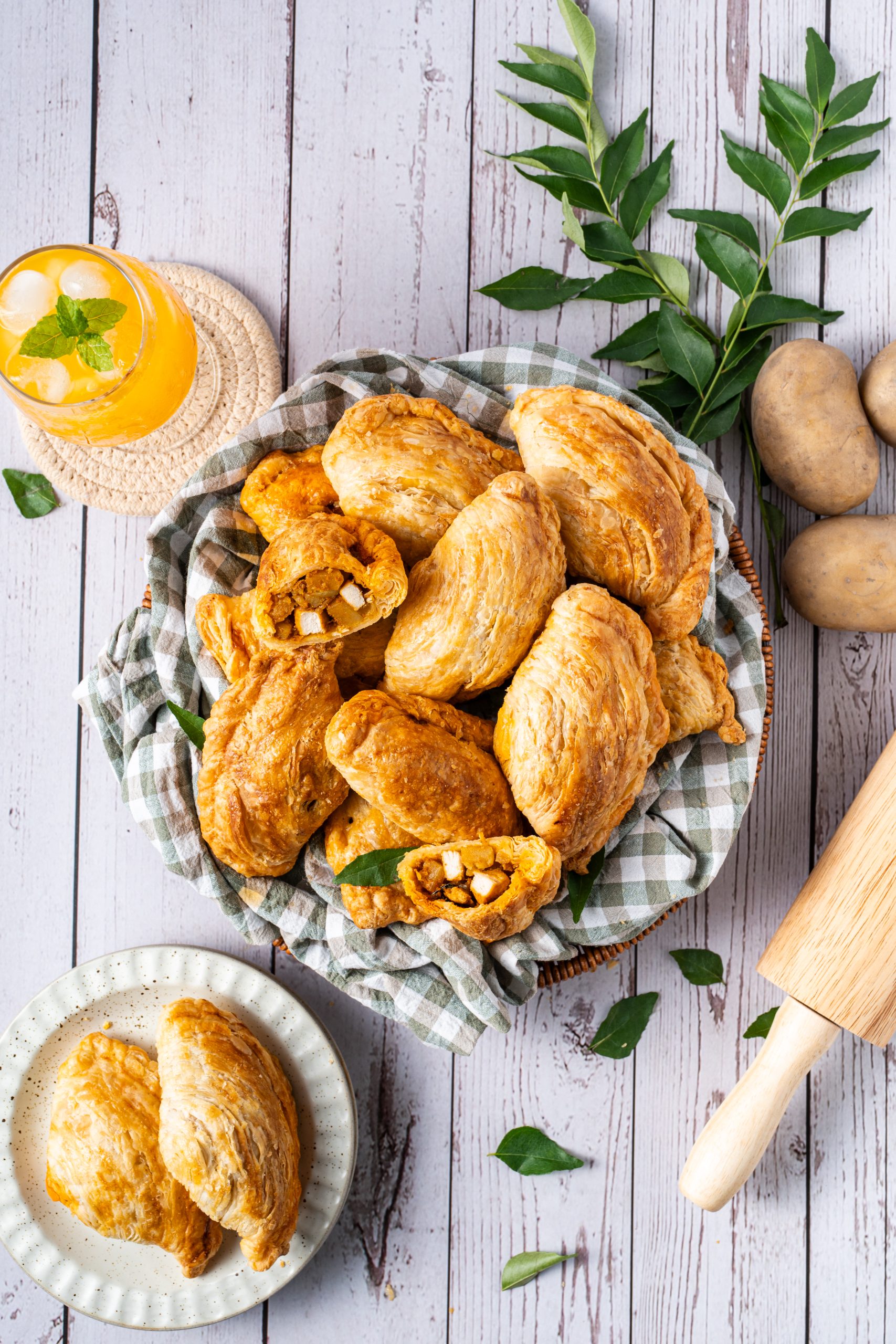 A basket of golden, flaky curry puffs filled with spiced potato and chicken, displayed on a checkered cloth. One puff is cut open to show the savory filling. The scene includes a glass of orange drink with ice, fresh curry leaves, and whole potatoes on a rustic wooden surface.