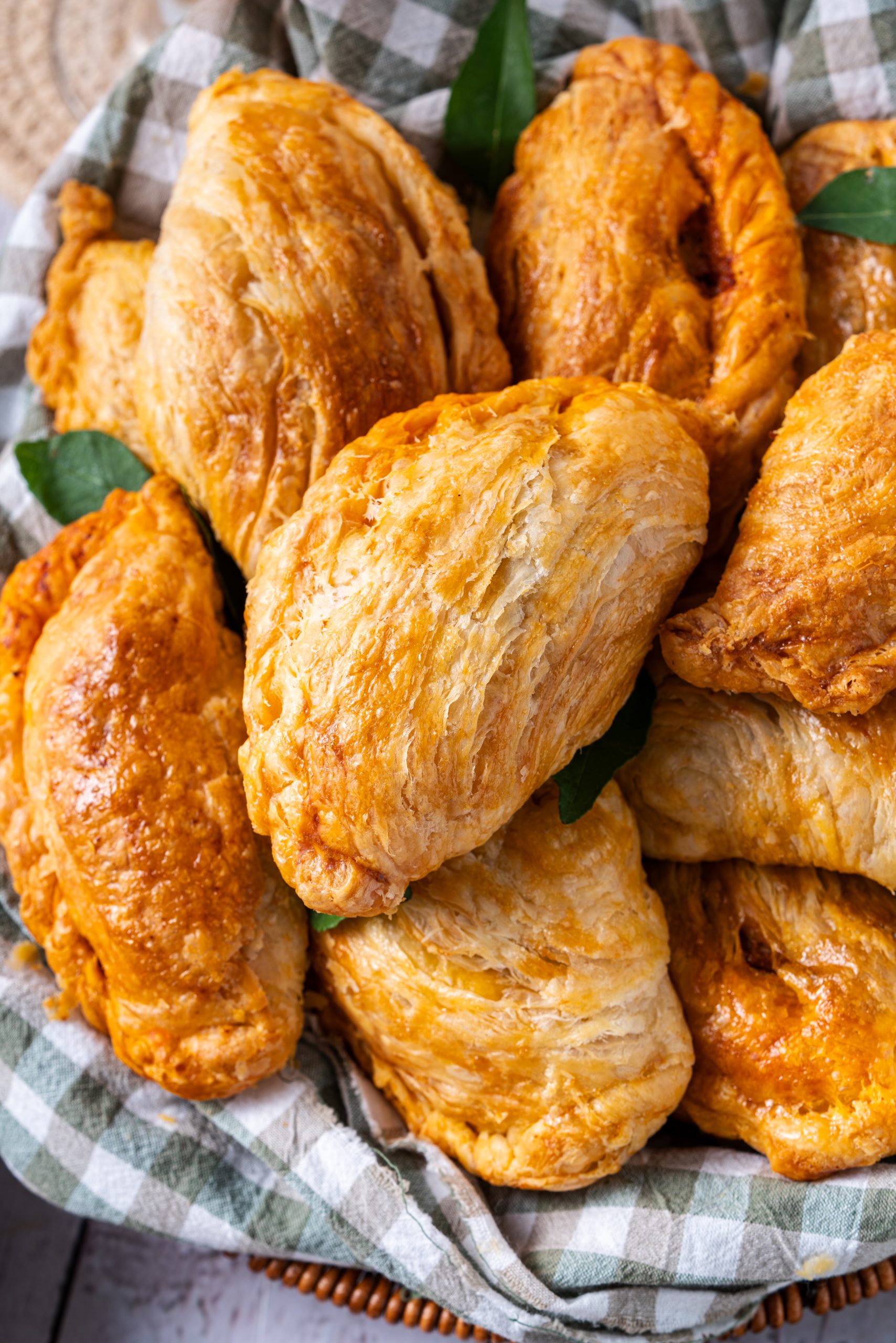 A close-up of freshly baked curry puffs with beautifully golden, flaky layers piled in a basket lined with checkered cloth. The crispy pastry texture is highlighted, showing the rich, buttery sheen and inviting handmade look.
