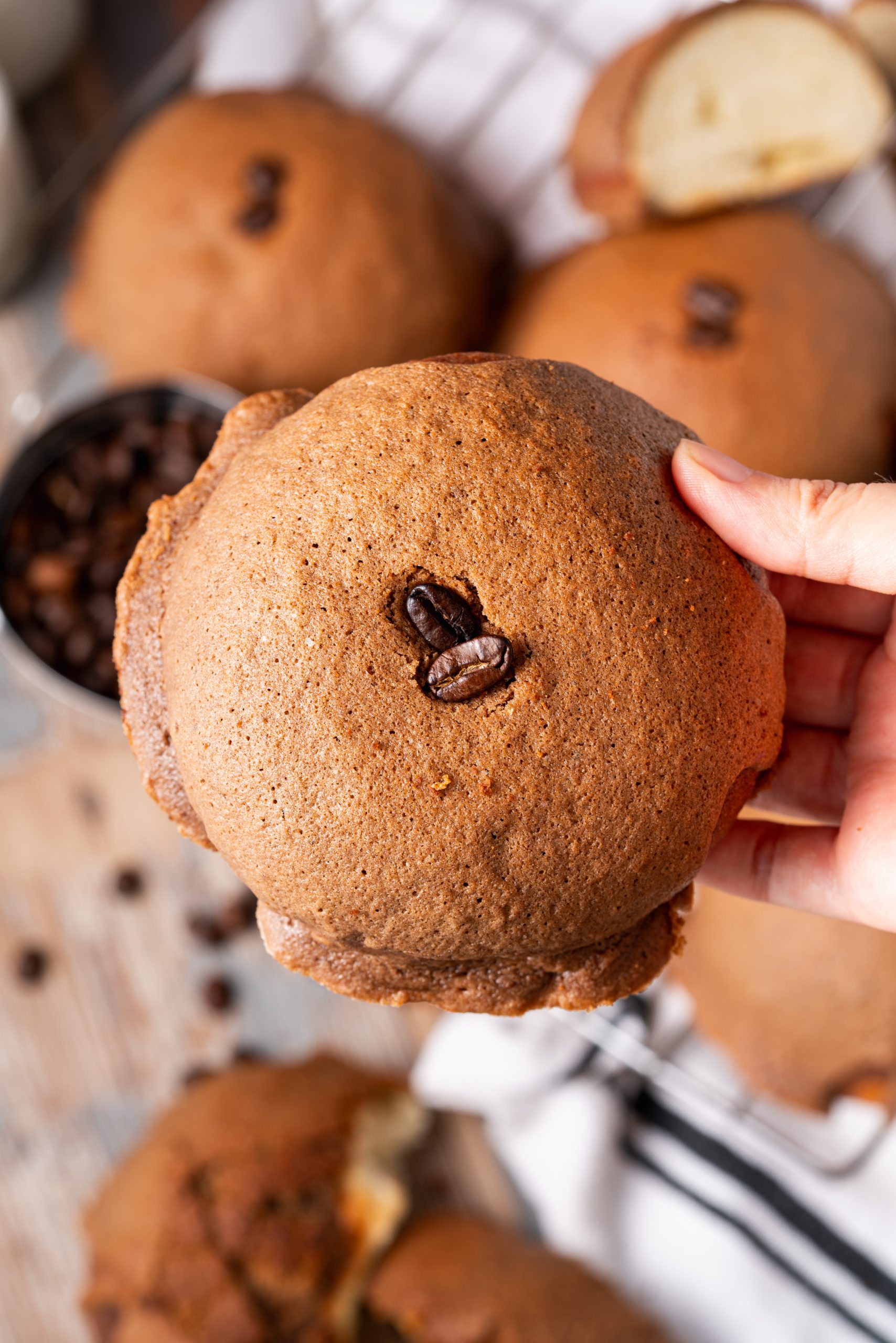 Hand holding a freshly baked Pappa Roti bun with visible coffee beans on top, showing its perfectly browned crust.