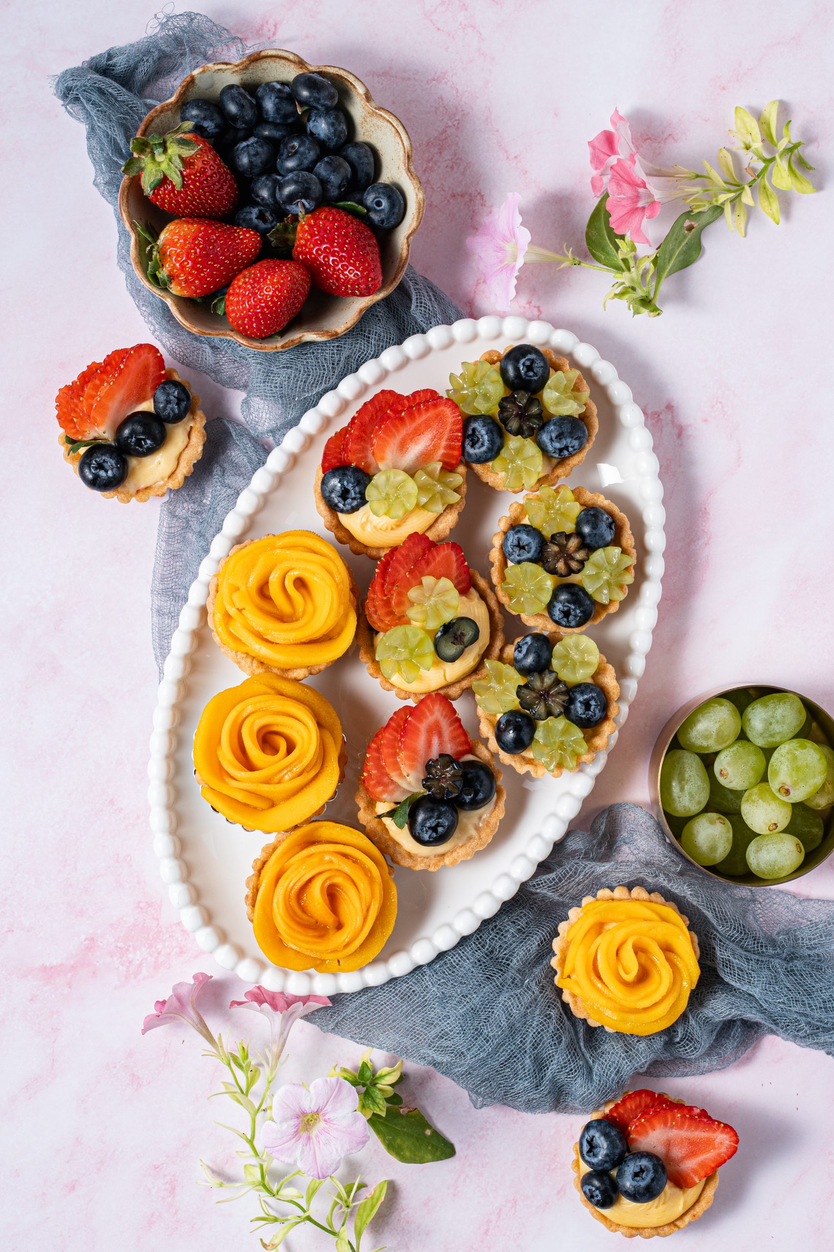 A bright and colorful assortment of mini fruit tartlets filled with custard and topped with fresh strawberries, blueberries, green grapes, and mango roses, arranged on a white platter over a soft pink background with flowers