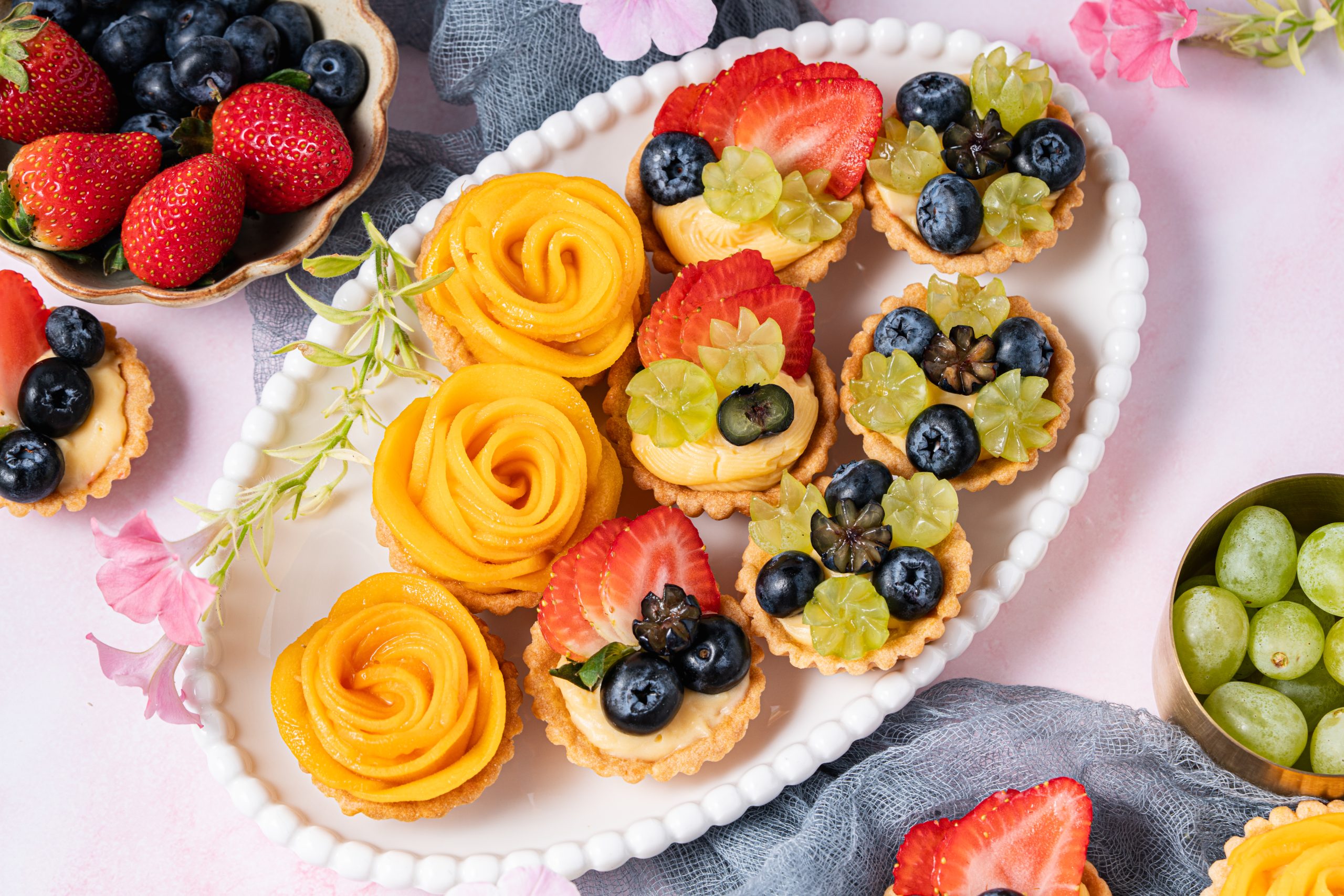 A plate of colorful mini fruit tartlets filled with creamy custard, topped with sliced strawberries, blueberries, green grapes, and mango rose spirals, surrounded by fresh fruit and flowers on a soft pink background.