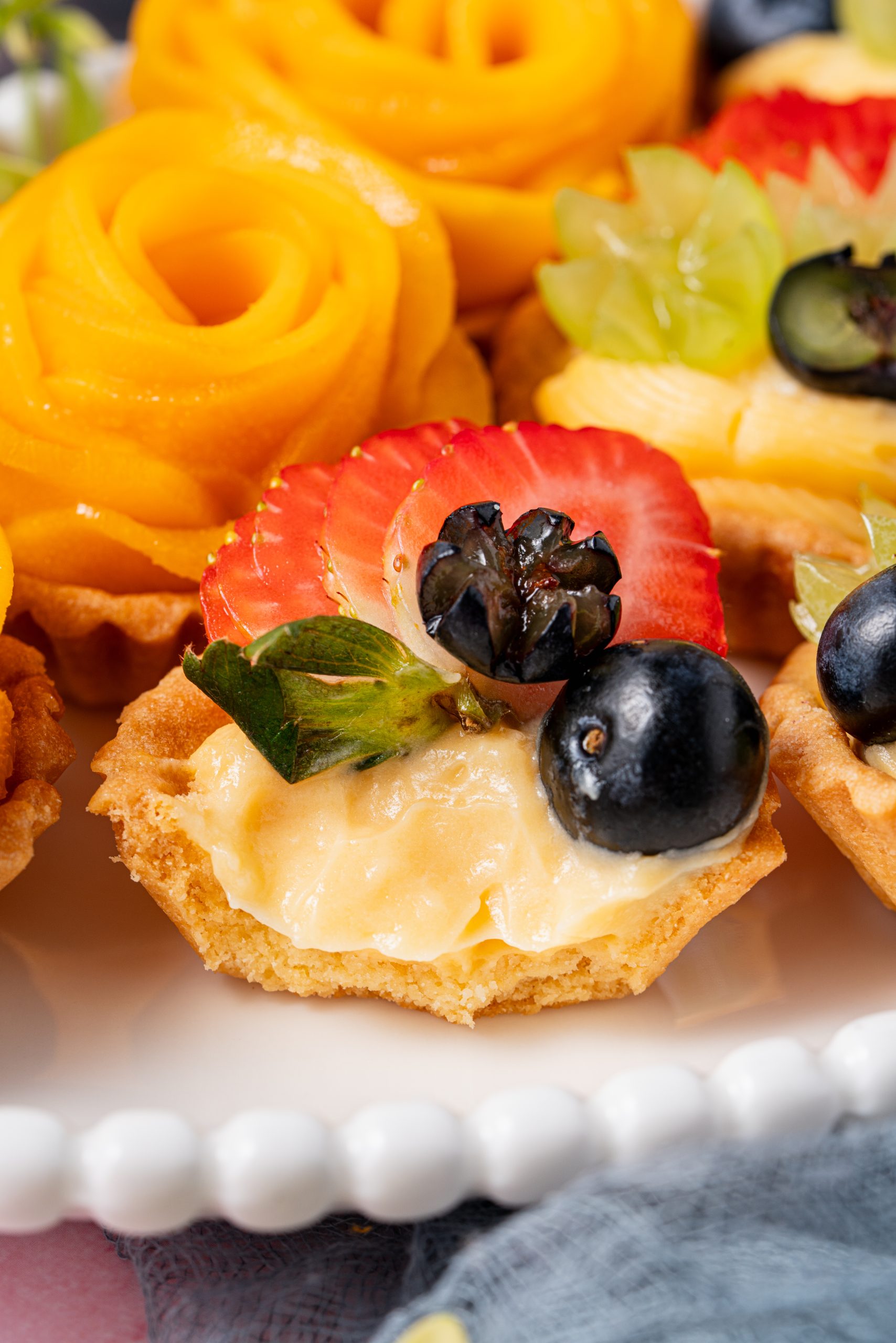 Close-up of a freshly baked fruit tartlet topped with strawberries, blueberries, and creamy custard, with mango rose tartlets in the background on a white serving plate.
