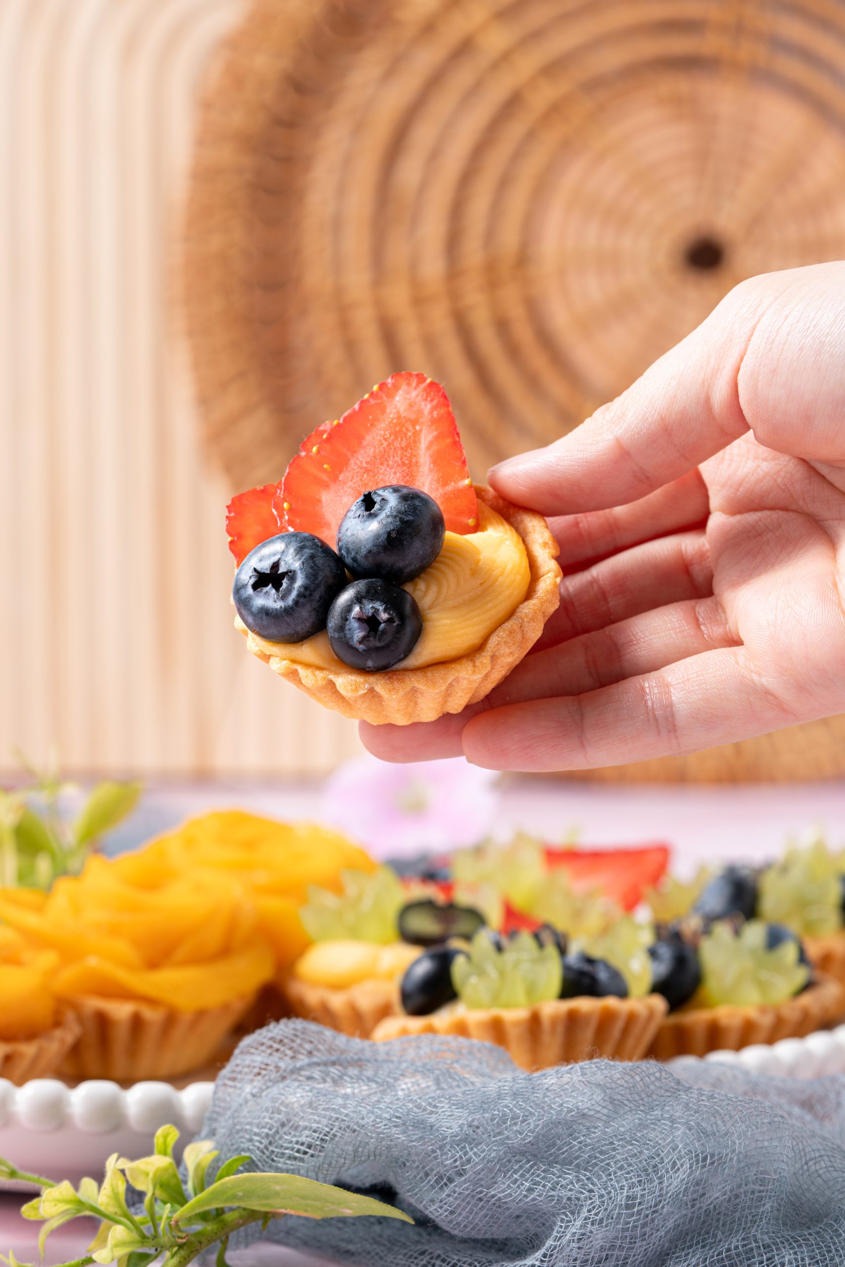 A hand holding a mini fruit tartlet topped with sliced strawberries and plump blueberries on creamy custard, with more tarts and fresh fruits blurred in the background.