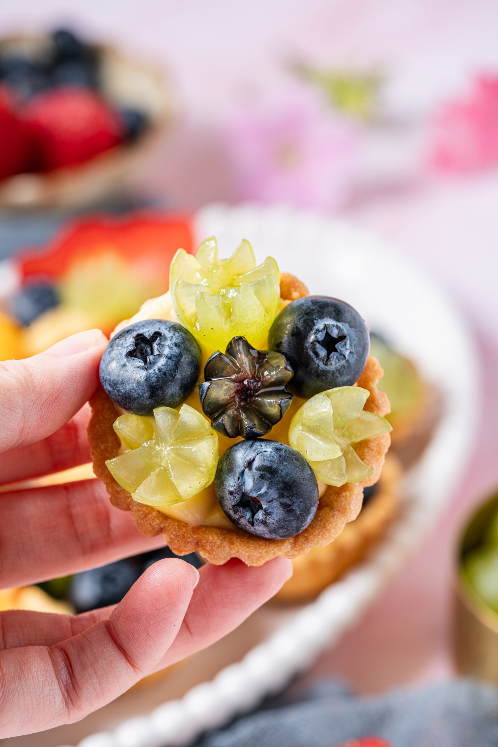 A hand holding a mini fruit tartlet topped with blueberries and green grapes over a pastel background.