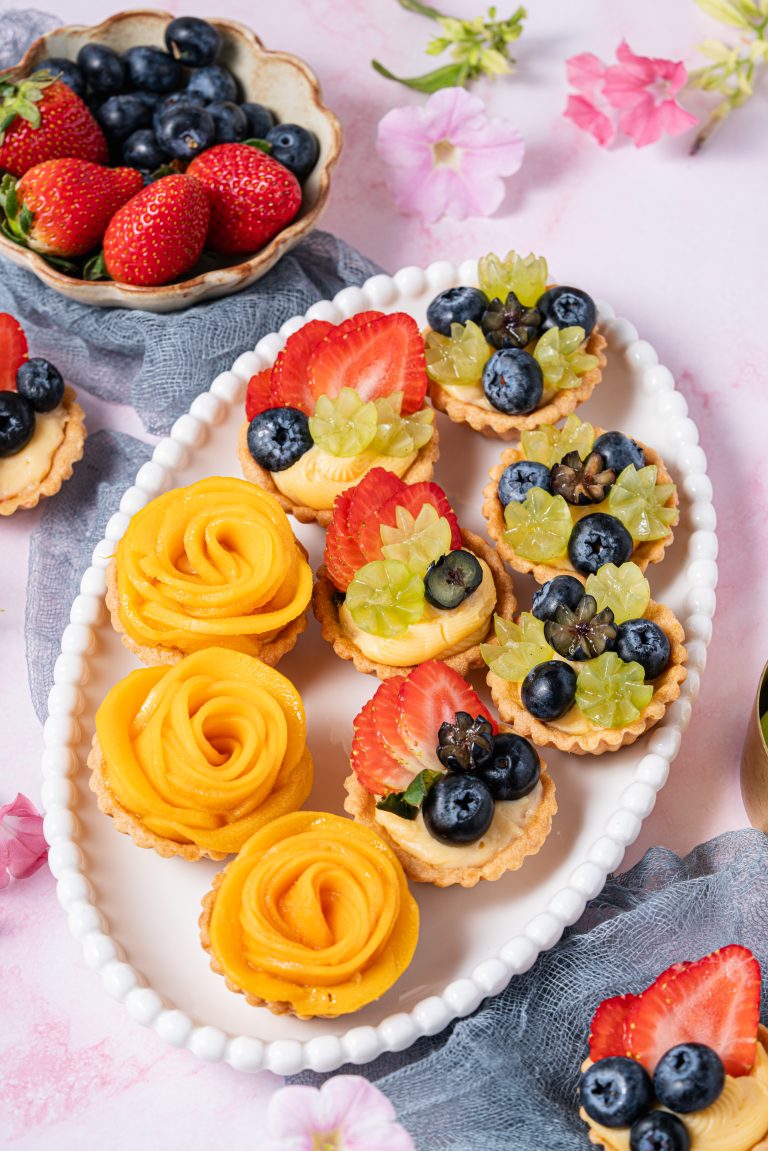 Overhead view of assorted mini fruit tartlets with mango, strawberry, and blueberry toppings, arranged on a white plate with fresh fruits in the background.