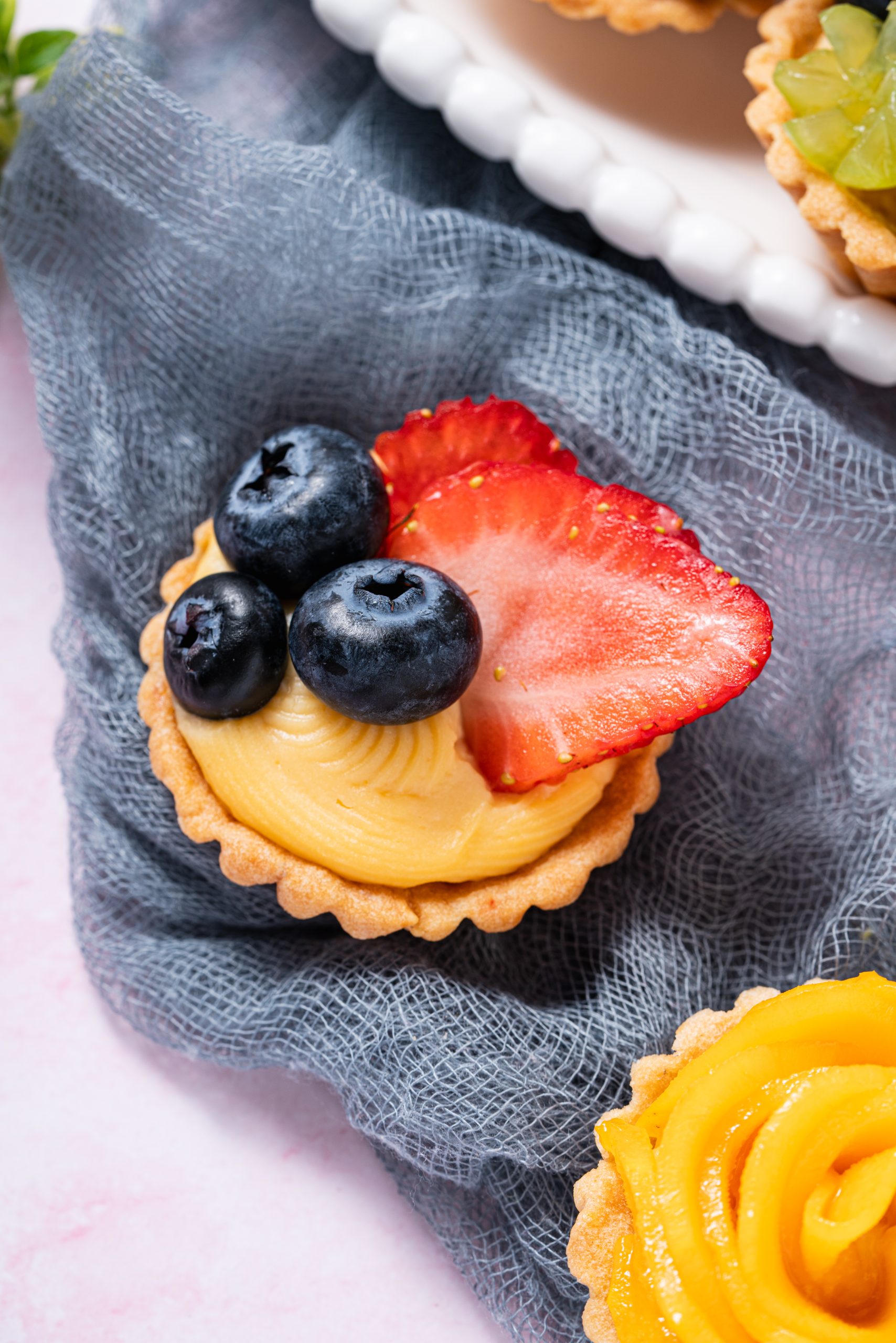 Single mini fruit tartlet topped with sliced strawberries and blueberries on a soft gray fabric background.