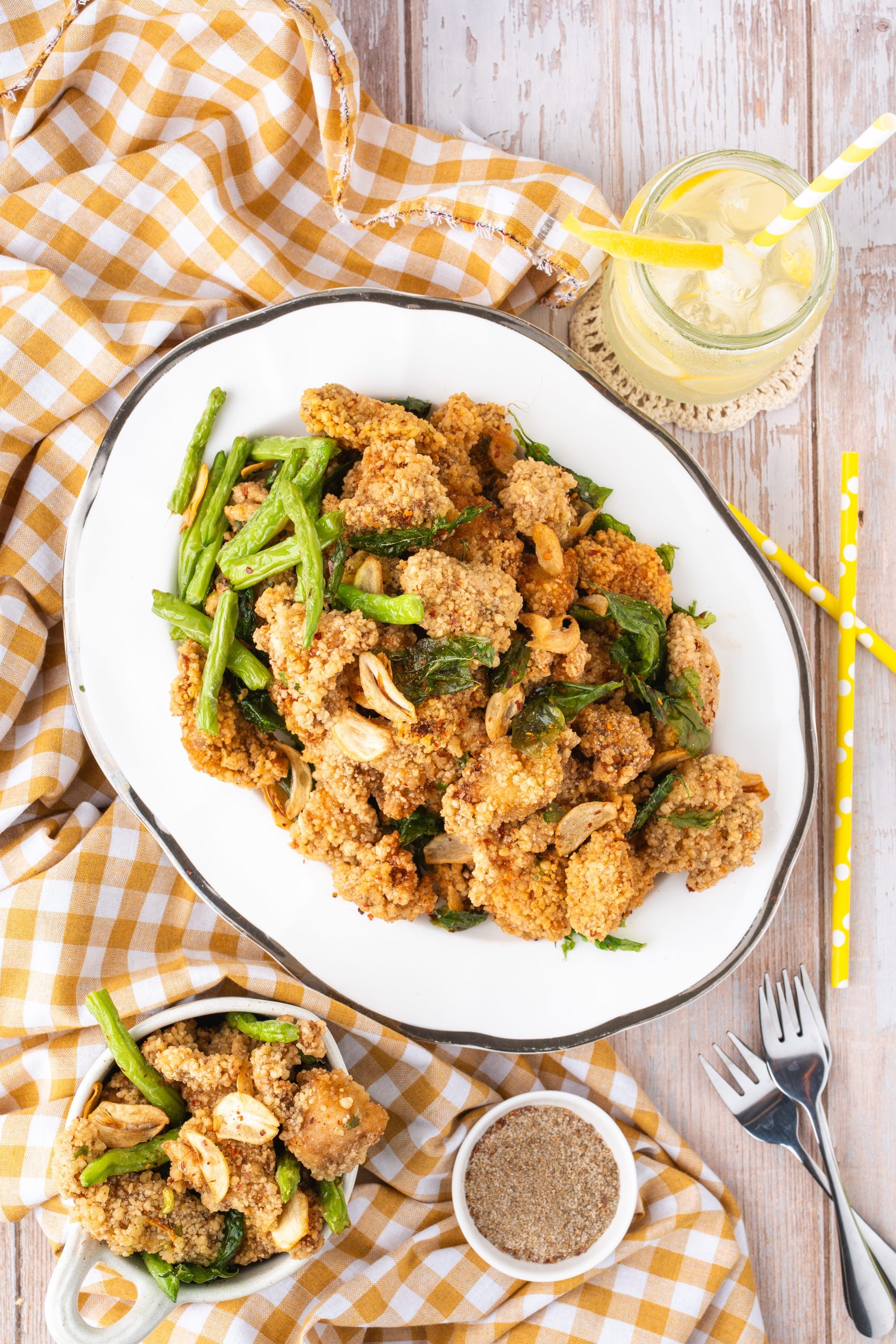 Overhead view of a plate of Taiwanese Popcorn Chicken garnished with fried basil and green chili peppers, served with dipping seasoning and a glass of lemon drink on a wooden table with a yellow checkered cloth.