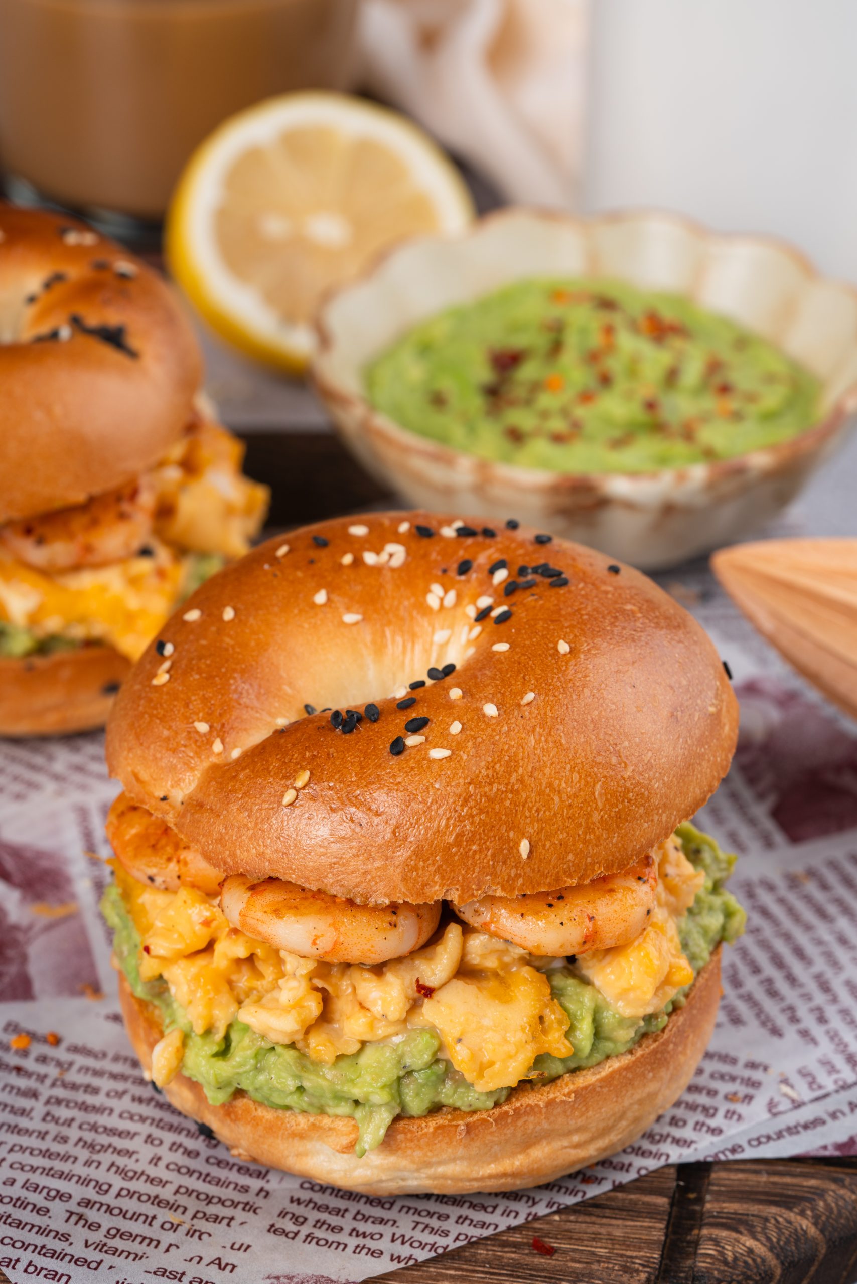 A delicious avocado shrimp bagel sandwich filled with creamy scrambled eggs, mashed avocado, and grilled shrimp, topped with sesame seeds and served on parchment paper.