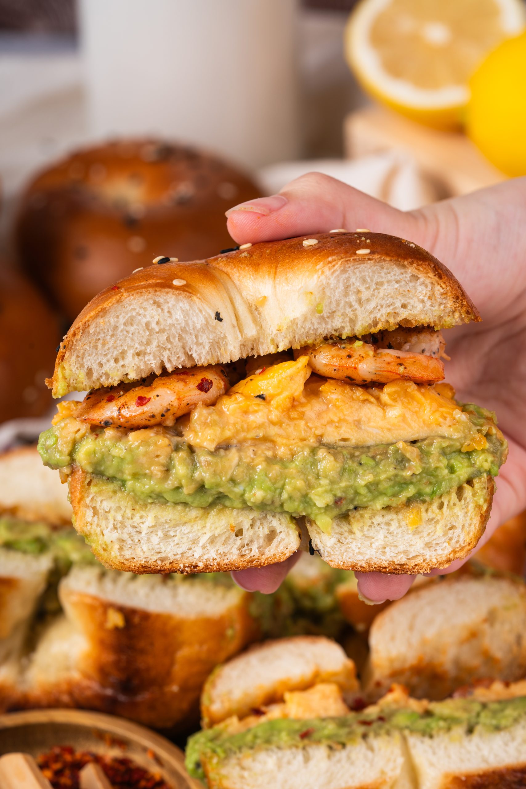 Hand holding half of a shrimp avocado bagel sandwich, displaying its rich layers of avocado, scrambled eggs, and shrimp filling against a background of fresh lemons and milk bottle.