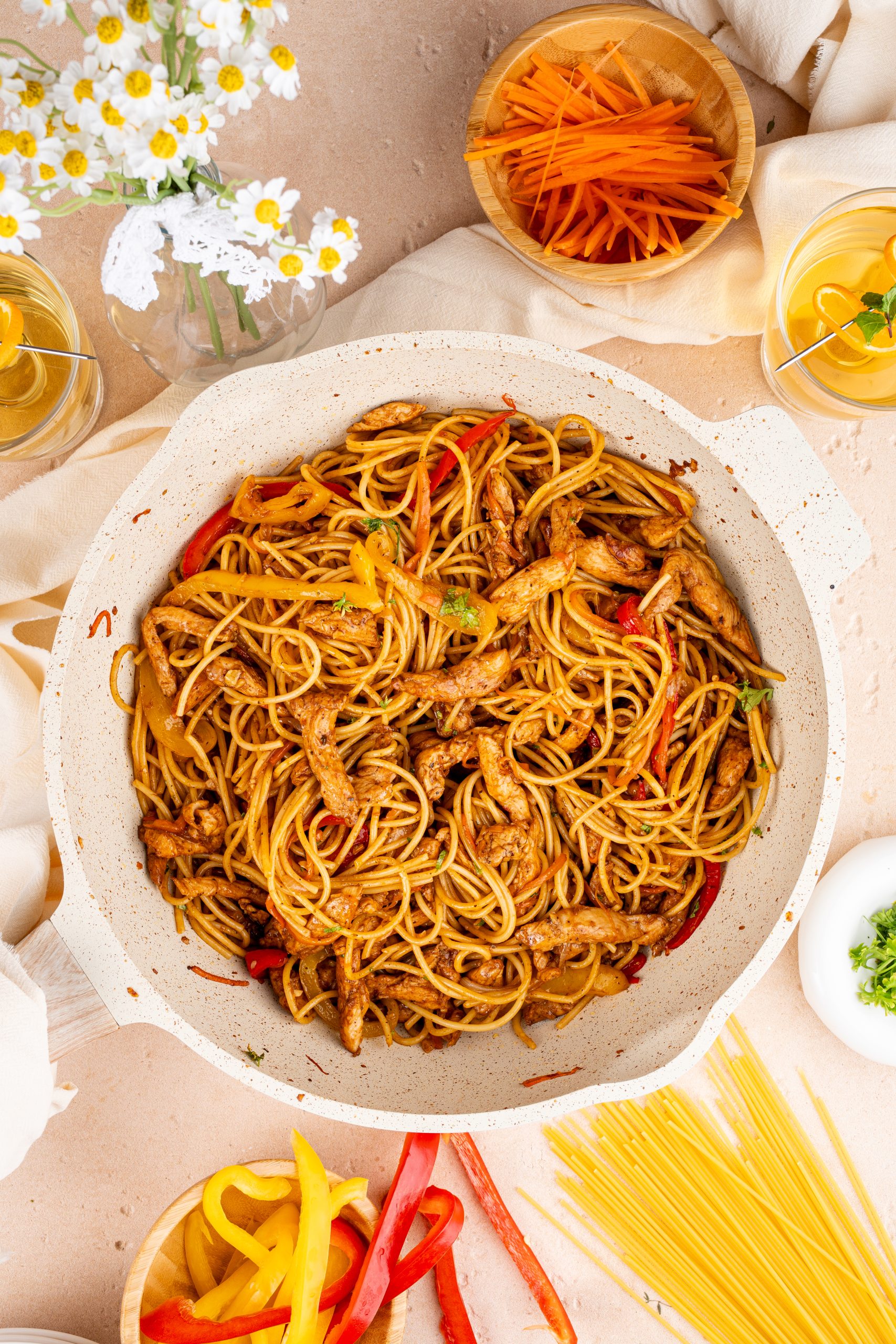 A pan of stir-fried pasta with chicken strips, red and yellow bell peppers, and carrots, coated in a savory sauce. The dish is surrounded by raw vegetables, spaghetti, and glasses of iced tea on a beige background.