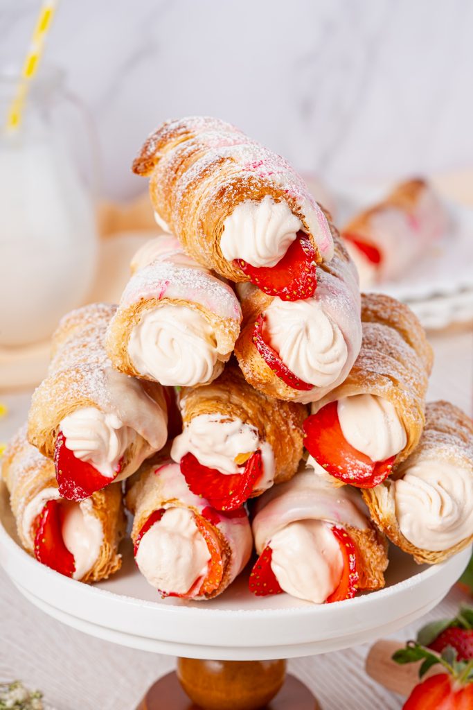 Close-up of freshly baked Cream Horns filled with smooth whipped cream and sliced strawberries, dusted with powdered sugar and stacked elegantly on a white cake stand.