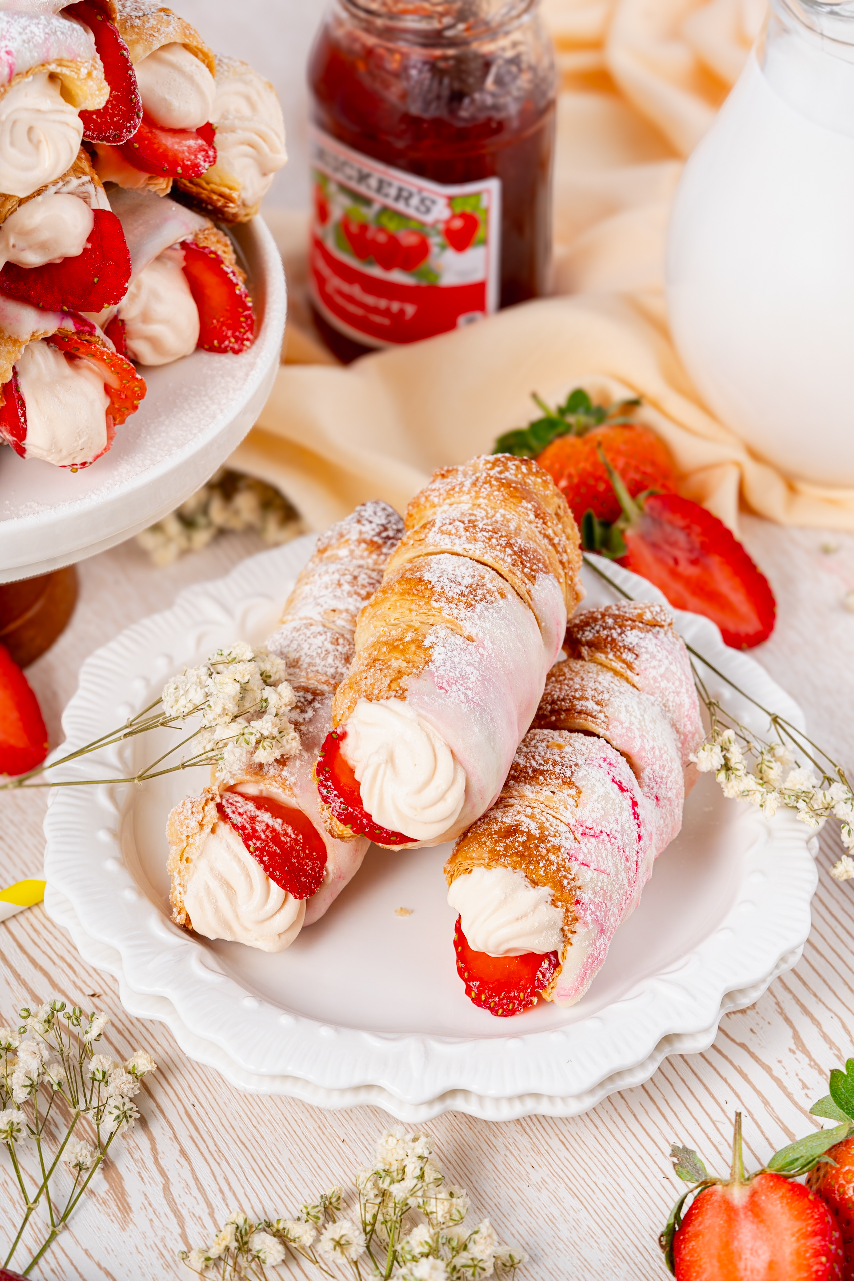 Three golden Cream Horns filled with whipped cream and fresh strawberries, dusted with powdered sugar and served on a white plate beside a jar of strawberry jam and a jug of milk.