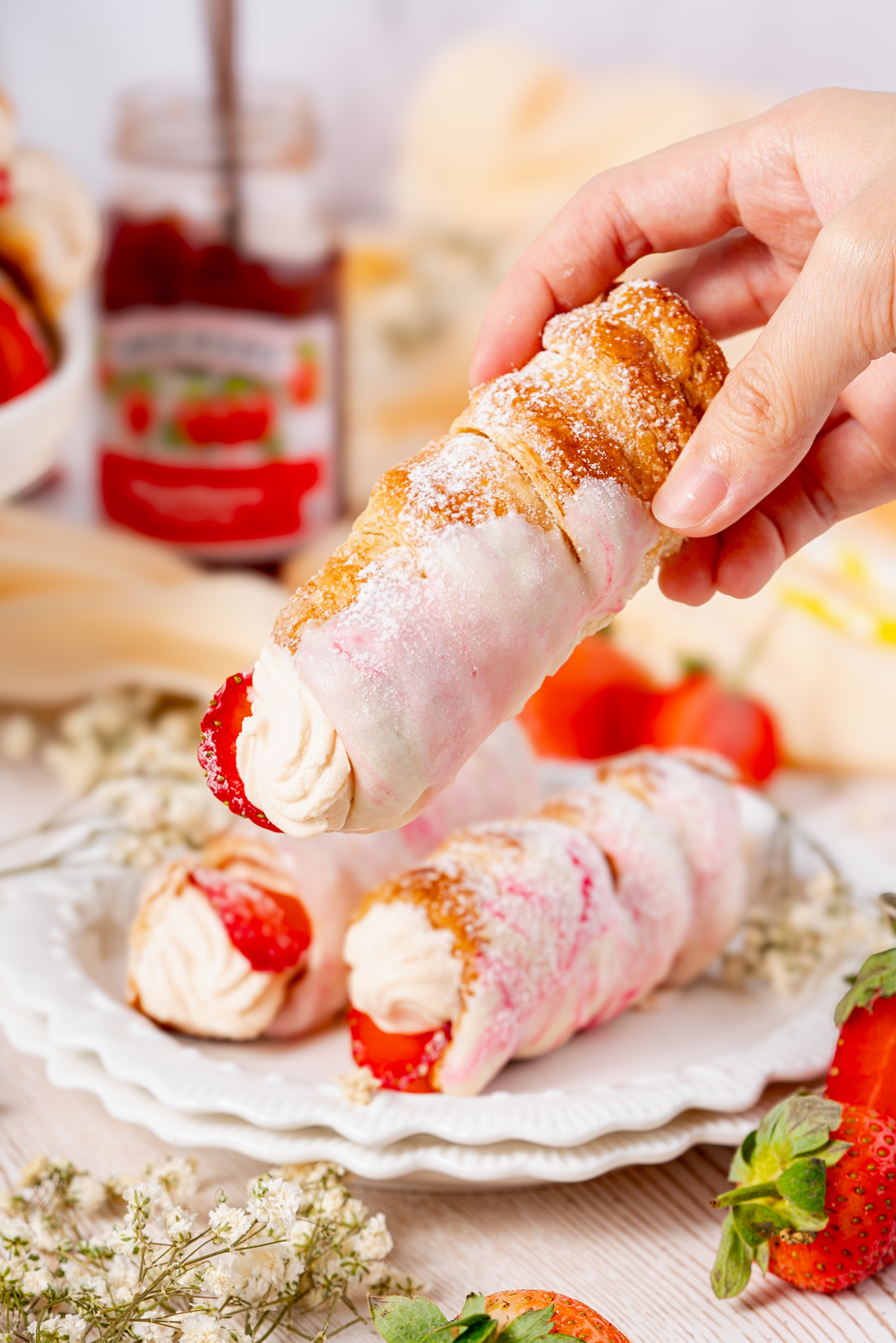 Hand holding a strawberry Cream Horn coated with pink glaze and powdered sugar, with a jar of strawberry jam in the background