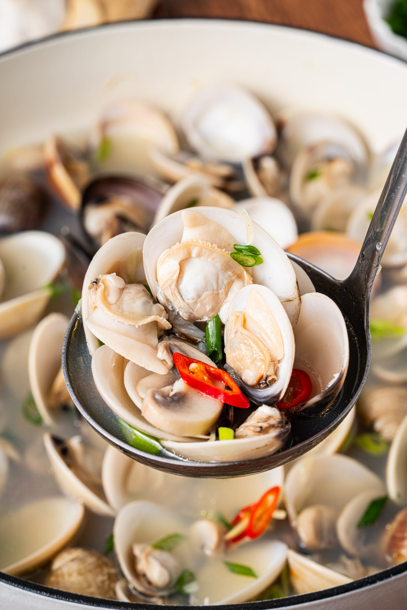 “Ladle lifting freshly cooked clams with mushrooms, green onions, and red chili peppers from a pot of soup.”