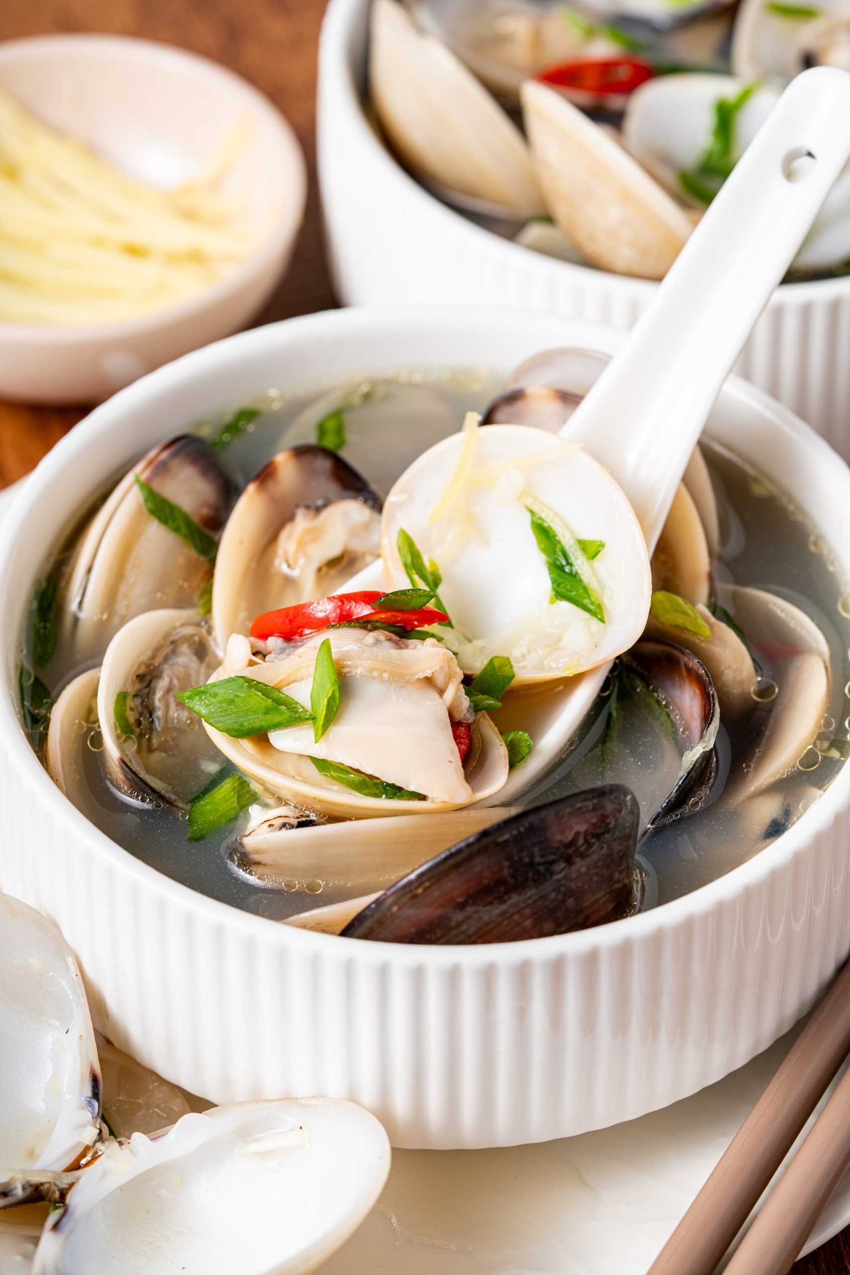 “Spoonful of clams, ginger, and green onions being lifted from a bowl of Asian clam soup.”