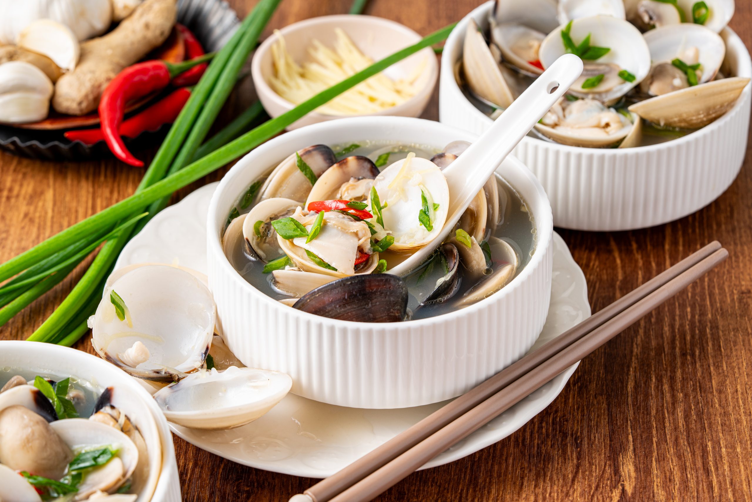 “Multiple bowls of clam soup with chili, ginger, and green onions arranged on a wooden table with chopsticks.