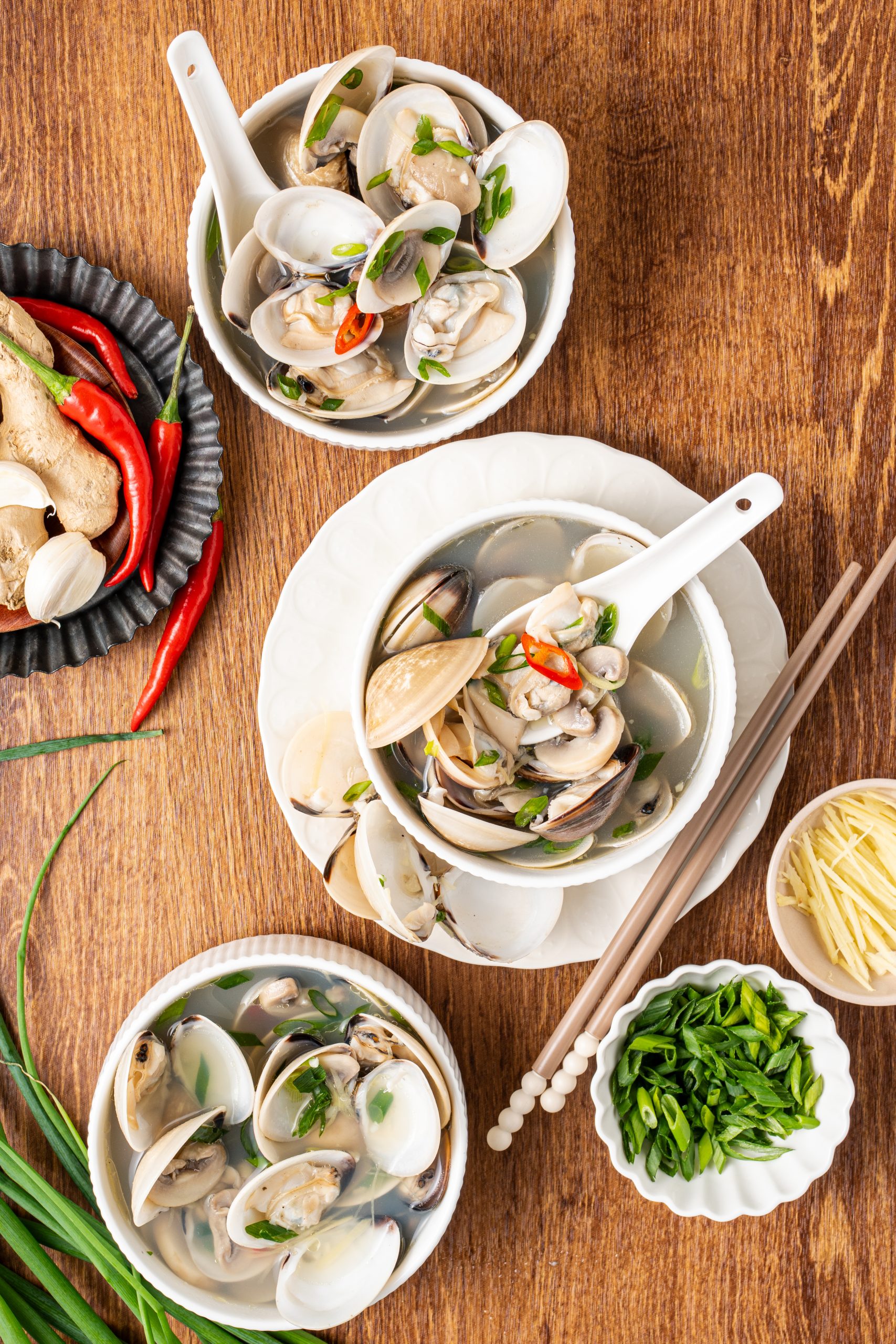 Top view of three bowls of Asian clam soup garnished with green onions and chili peppers on a wooden table.”