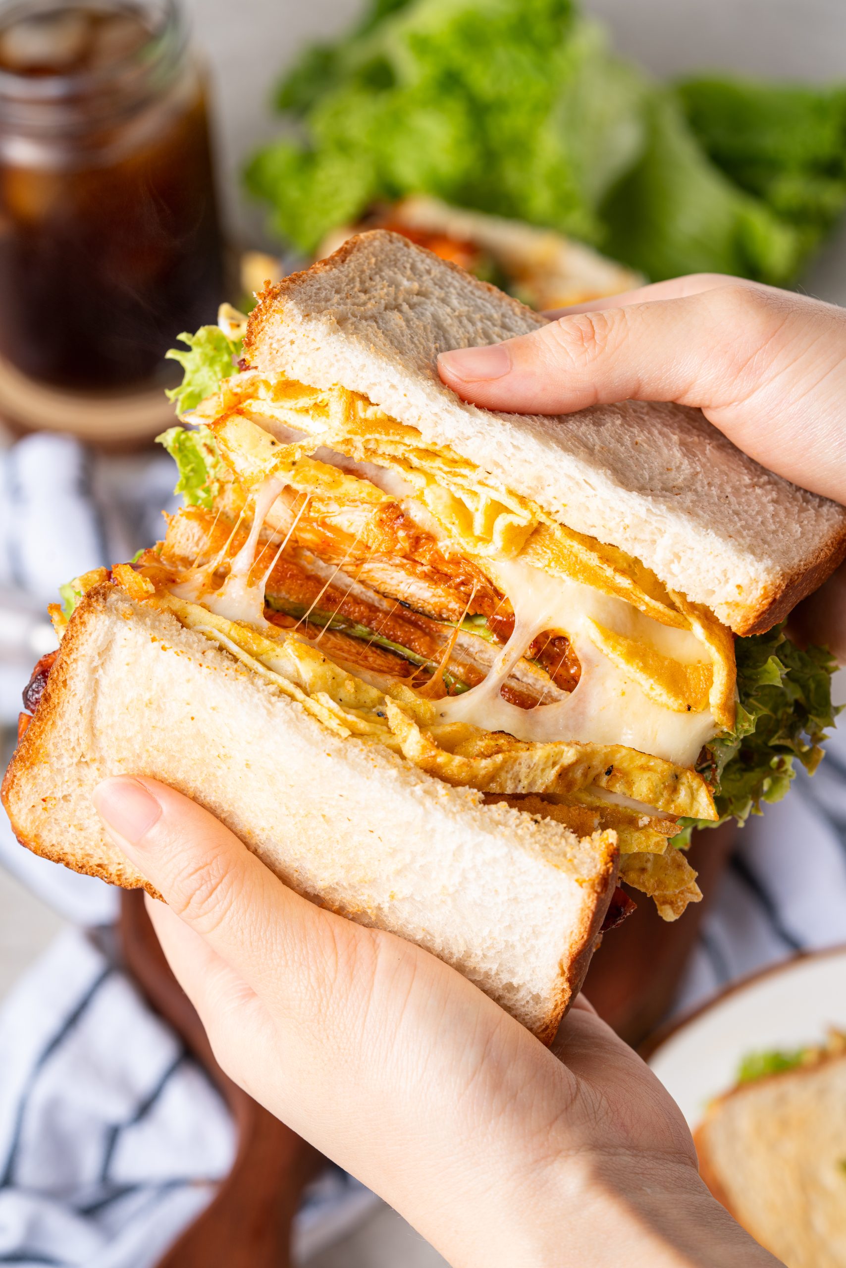 Hands pulling apart a gochujang chicken sandwich, showing melty cheese stretching between the sandwich layers, with chicken, lettuce, and egg strips inside.