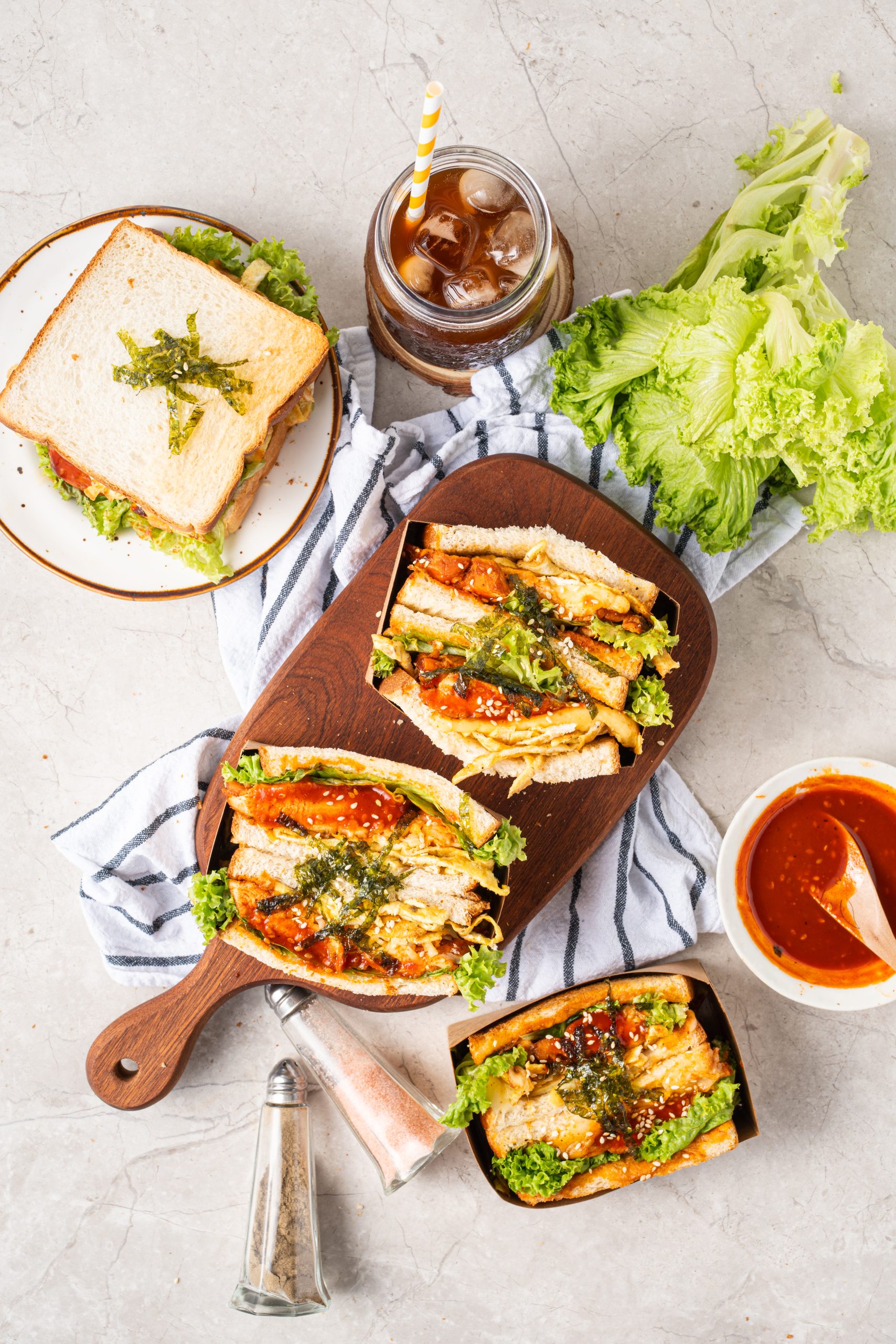 Top-down view of a sandwich and drink setup, showing multiple gochujang chicken sandwiches arranged on a wooden serving board with lettuce and iced tea.