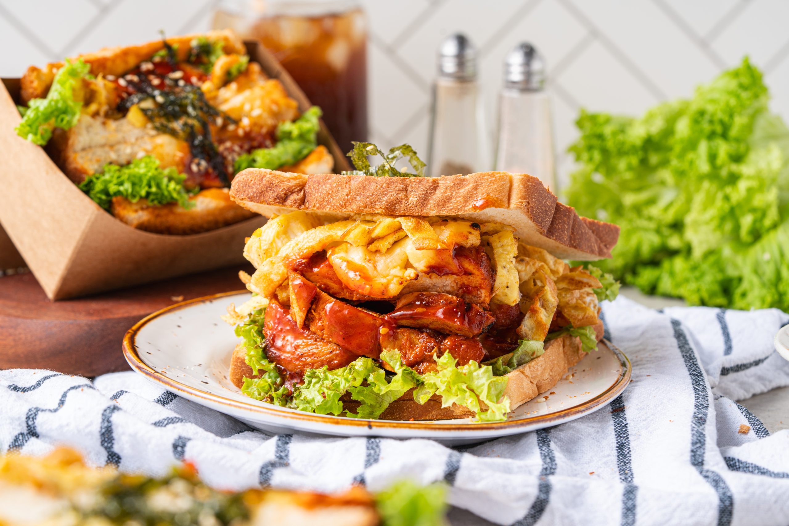 Gochujang chicken sandwich on a plate with iced tea and salt and pepper shakers in the background. The sandwich is layered with lettuce, chicken, egg strips, and sauce.