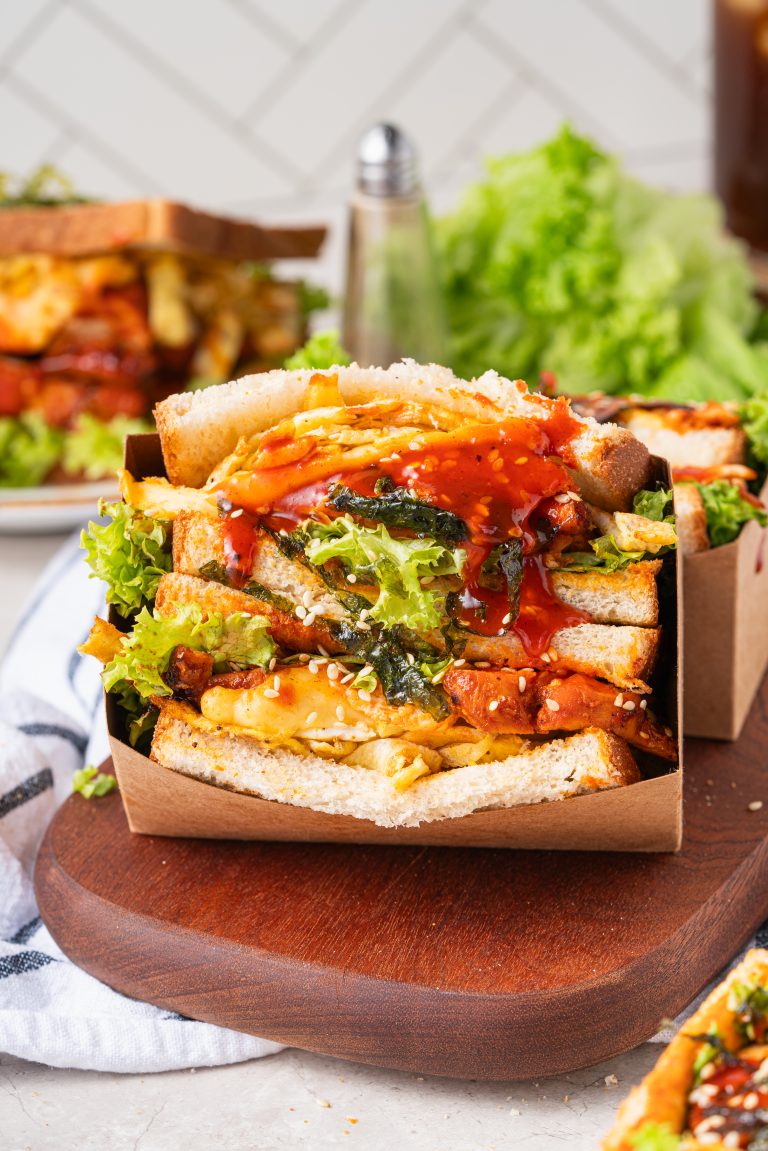 Close-up of a stacked gochujang chicken sandwich in a cardboard holder, filled with lettuce, grilled chicken, egg strips, and topped with sesame seeds and gochujang sauce.
