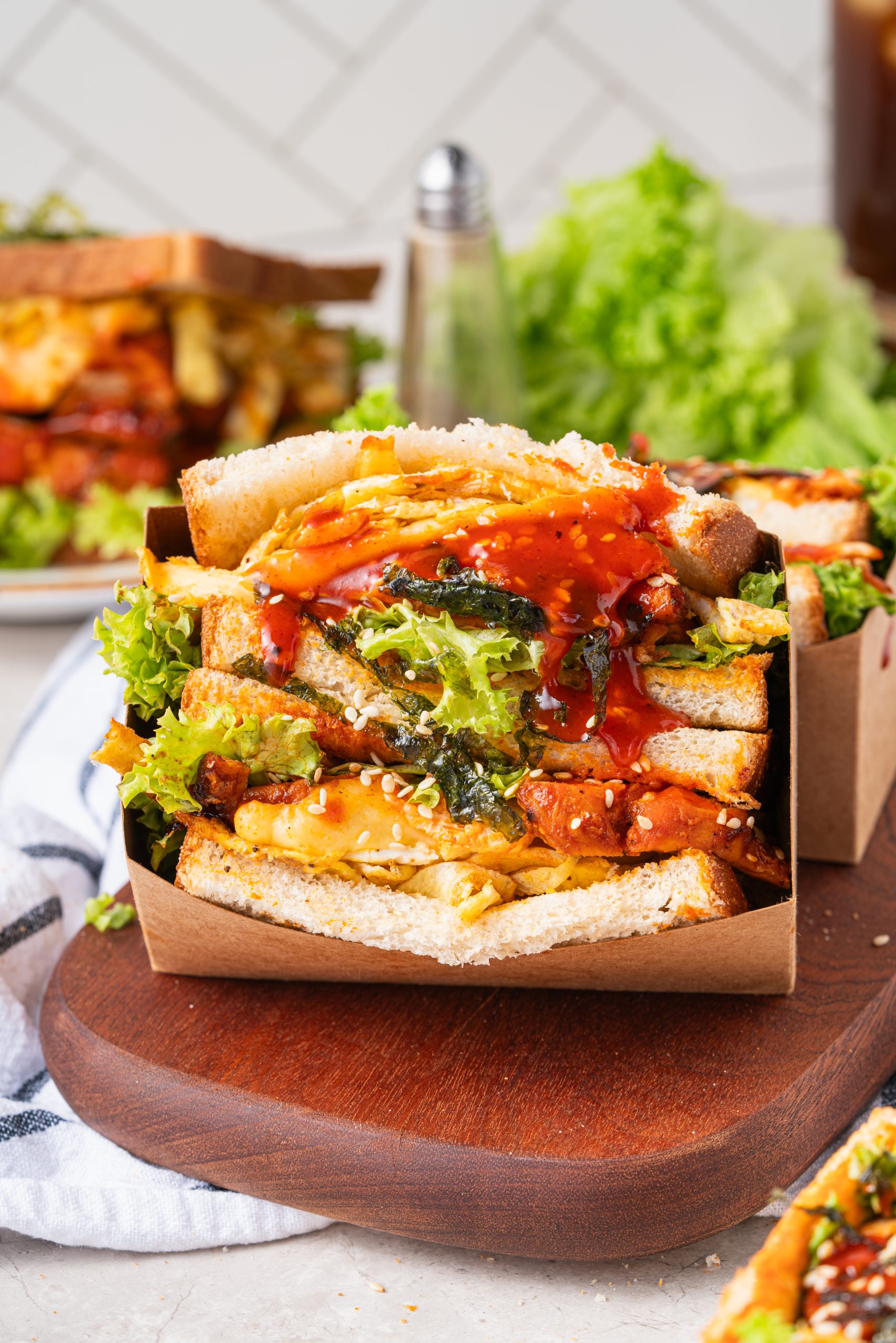 Close-up of a stacked gochujang chicken sandwich in a cardboard holder, filled with lettuce, grilled chicken, egg strips, and topped with sesame seeds and gochujang sauce.