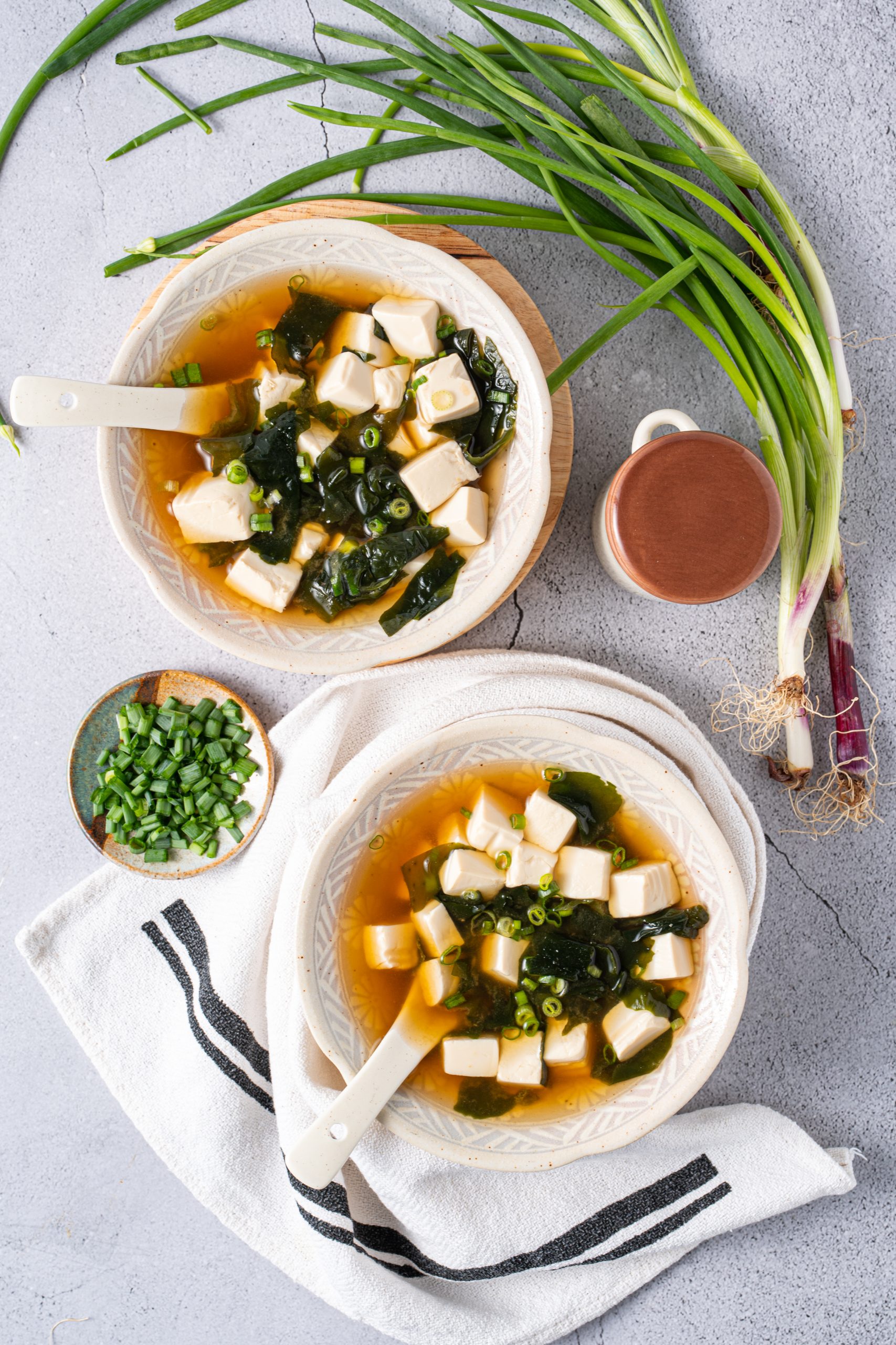 Two bowls of Japanese miso soup with tofu and seaweed, styled with fresh green onions and a wooden cup on a light stone surface.